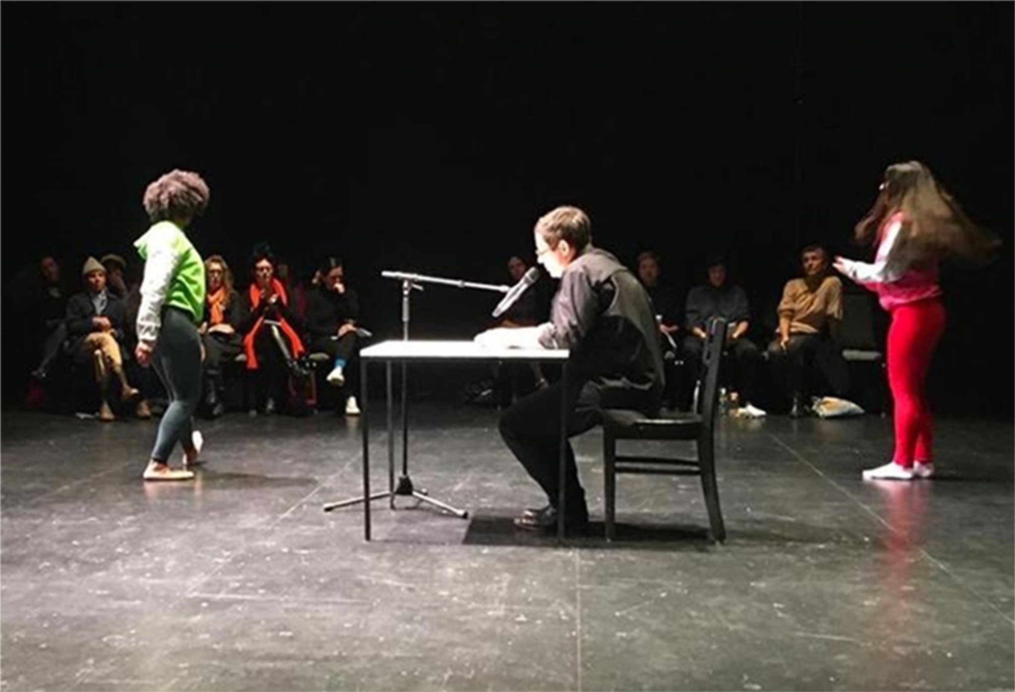 A photo of Andrea Geyer at a table speaking into a microphone. Geyer sits in a black box theater with performers on either side of the table and an audience seated in the background. 