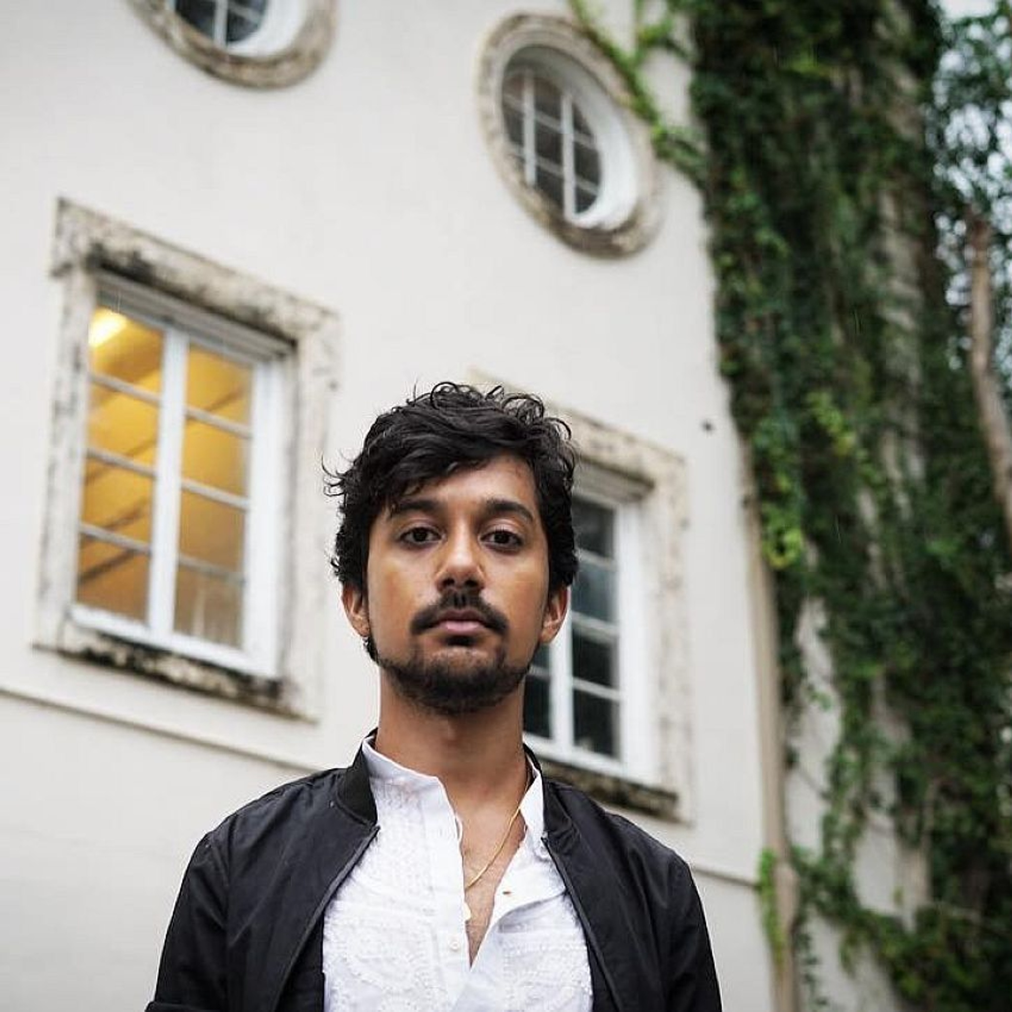 A portrait of artist Zain Alam. He is an Indian Pakistani person with wavy black hair parted over his forehead and a mustache. We see him from an angle slightly below, looking up at him with the façade of a house with windows in the background. He looks directly at us, eyes angled down slightly.