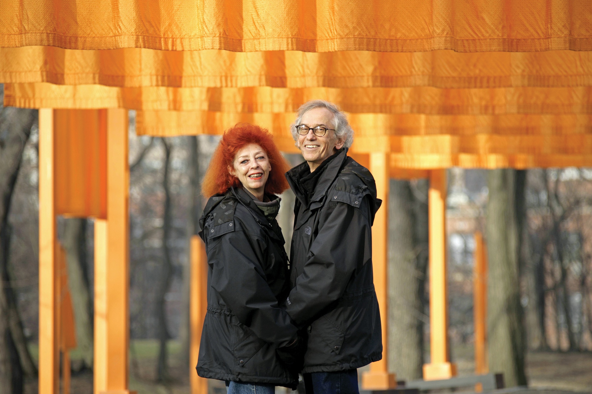 The artists Christo and Jeanne-Claude stand facing each other and holding hands in Central Park. Behind them hang the bright, saffron-colored fabric of their public art installation The Gates. 