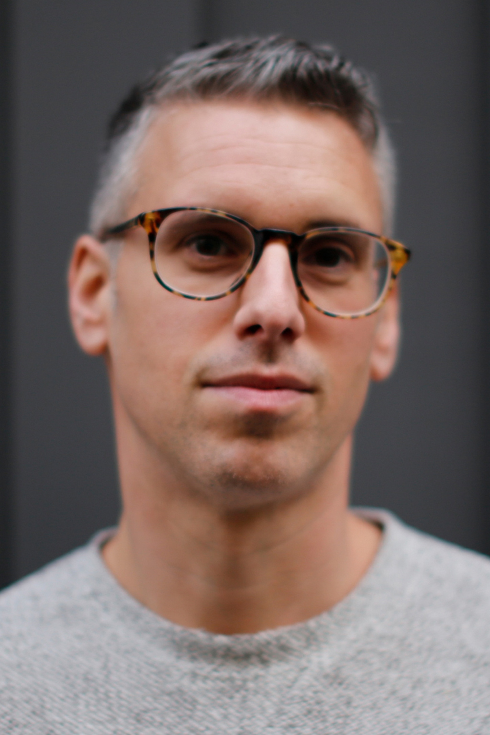 A portrait of graffiti artist Qu One seen from the shoulders up against a dark background. Qu One is a white man with short salt and pepper hair. He wears glasses looking intently, directly at us. He wears a gray tshirt.