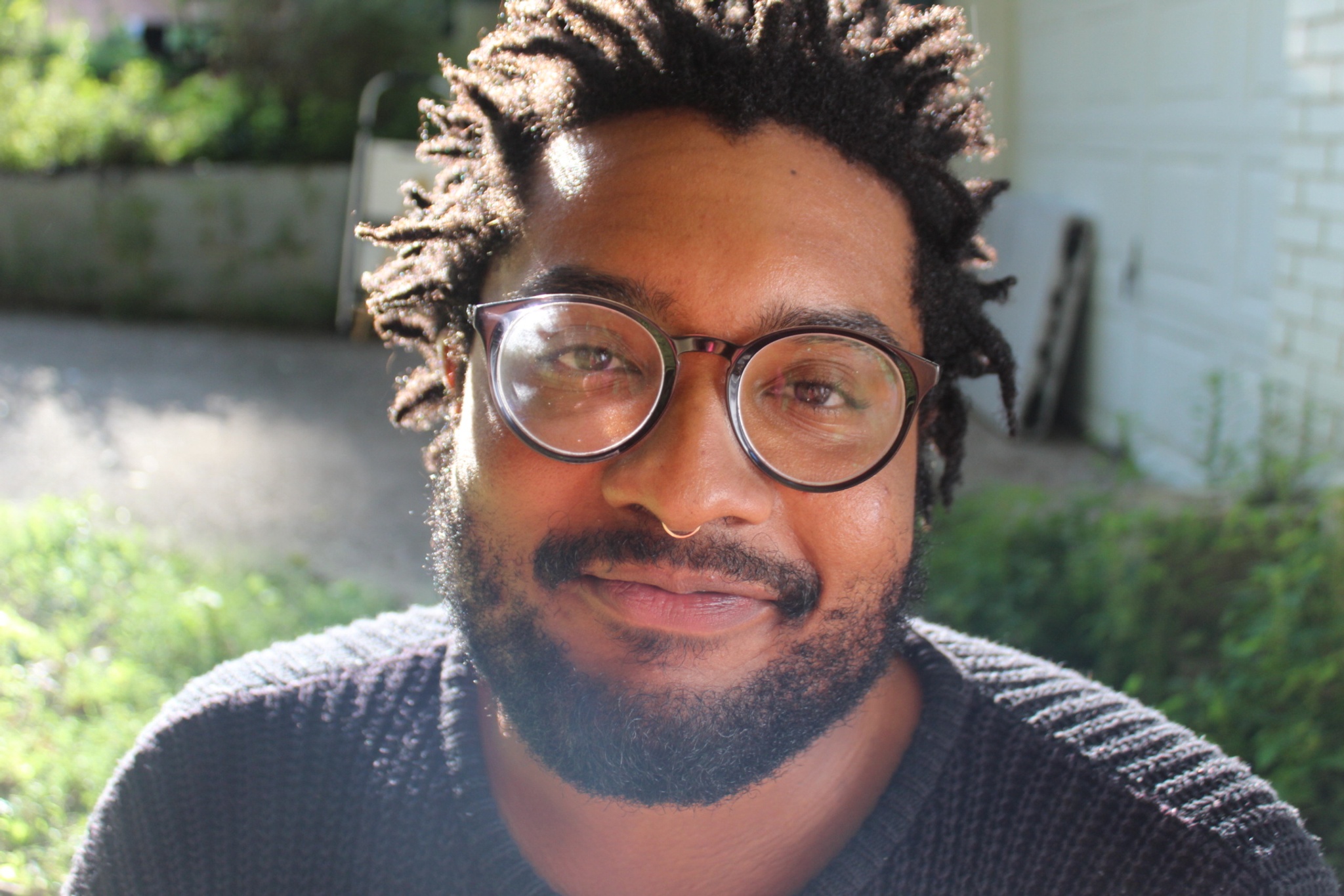 A photo of the artist Nazareth Hassan who has a beard and wears glasses, smiling against an outdoor background with grass and sunshine. 