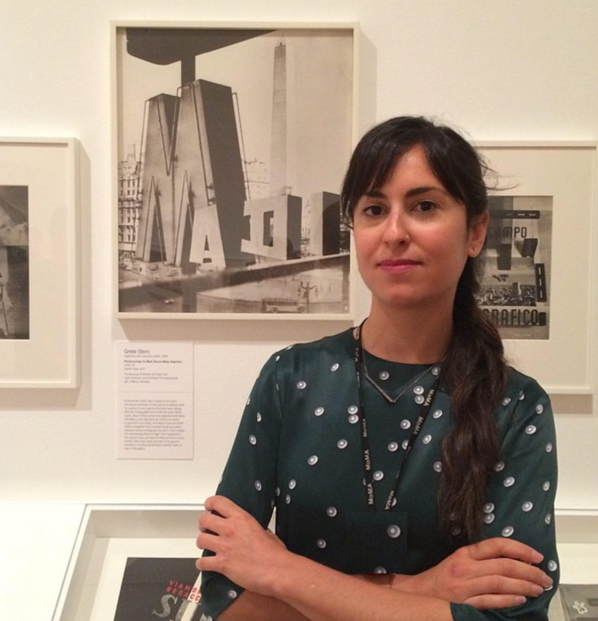 A photo of Karen Grimson standing with arms crossed in front of a museum display of photos by the photographer Grete Stern. 