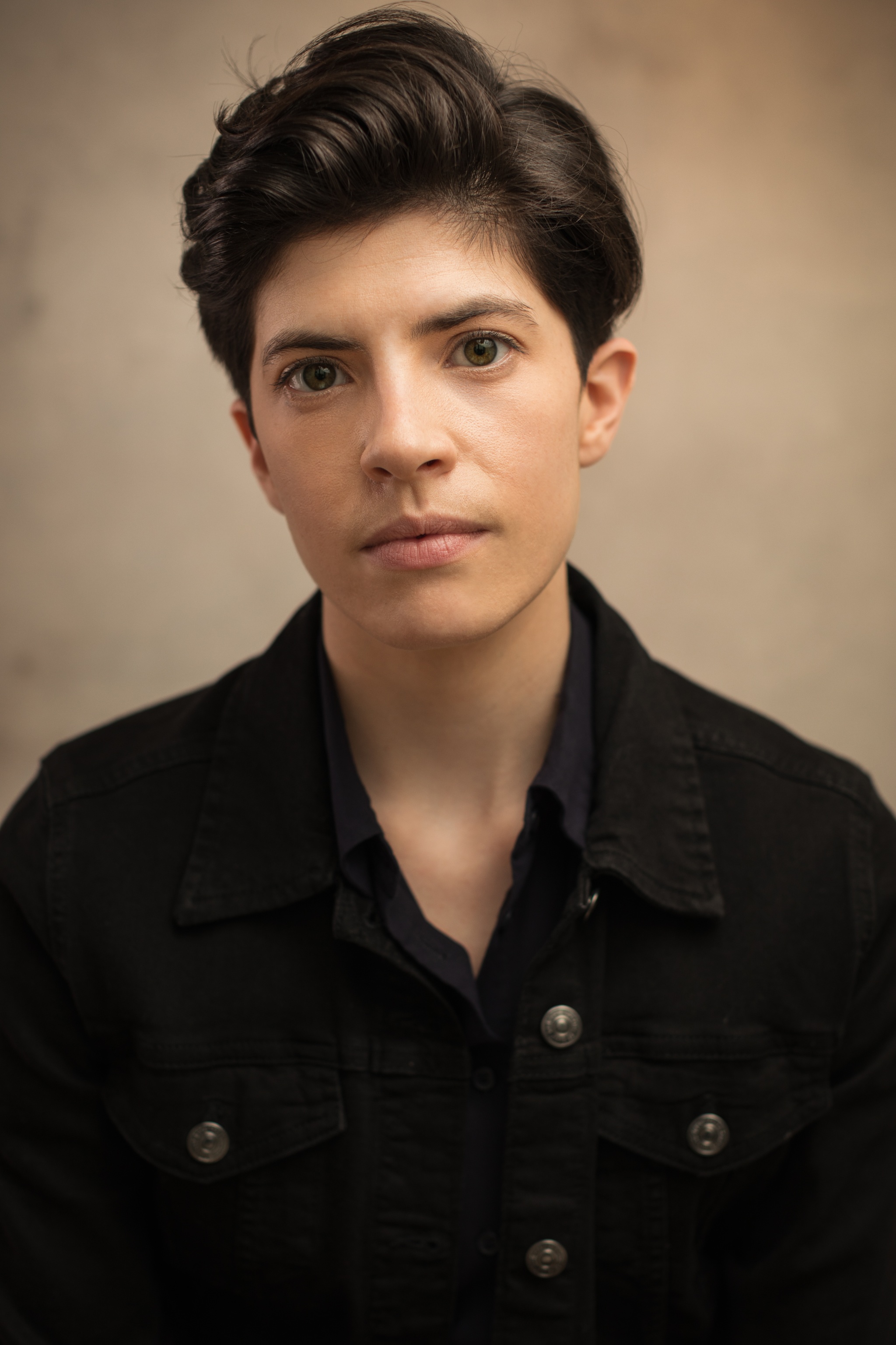 A headshot of actor Chloe Fenwick Brown, who looks intently at us. Chloe is white with short, dark brown hair and wears a black double breasted jacket. 