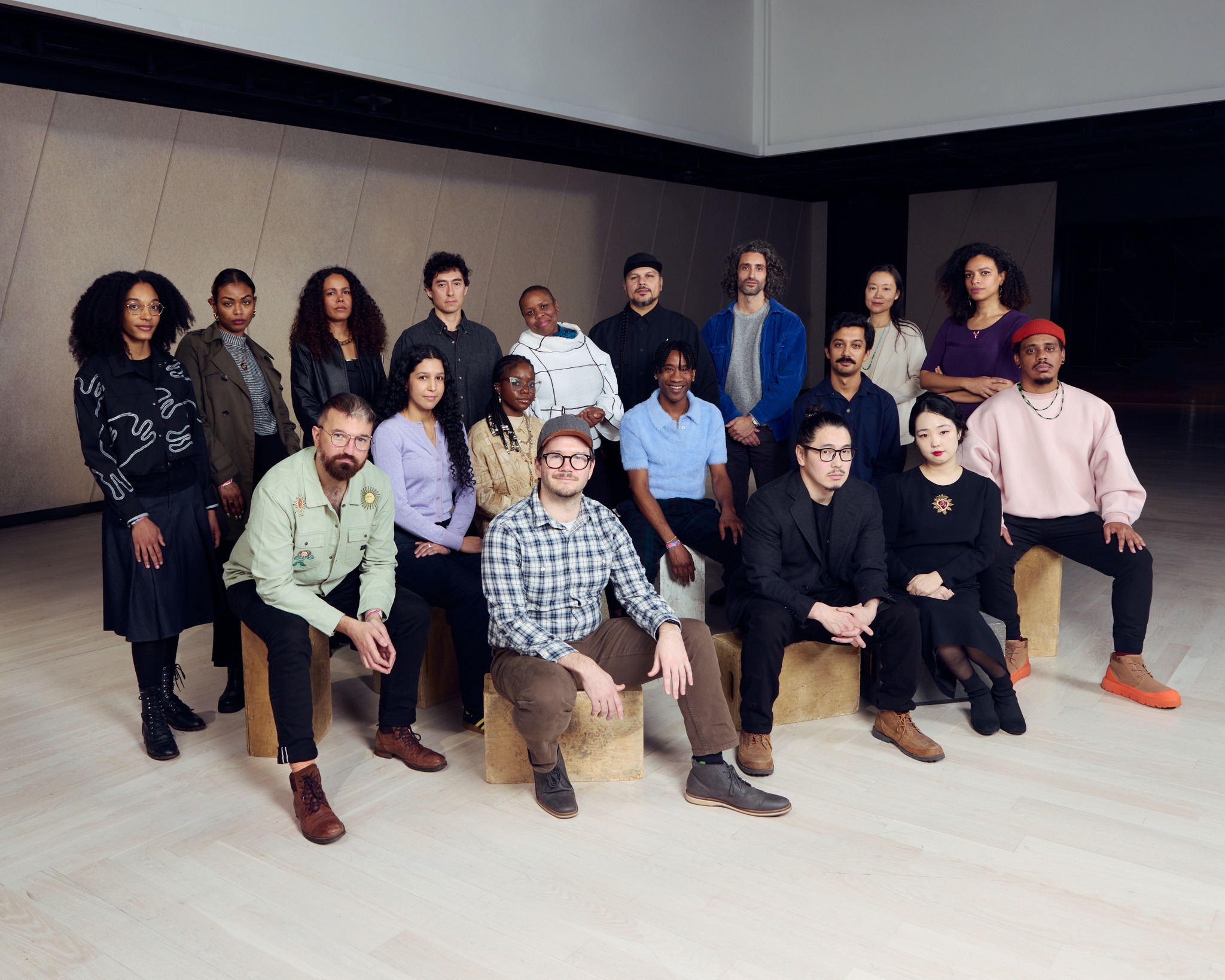 A group of artists pose for a photo in an empty event space. They are arranged in three rows, with the first two rows seated on crates and the back row standing behind them.