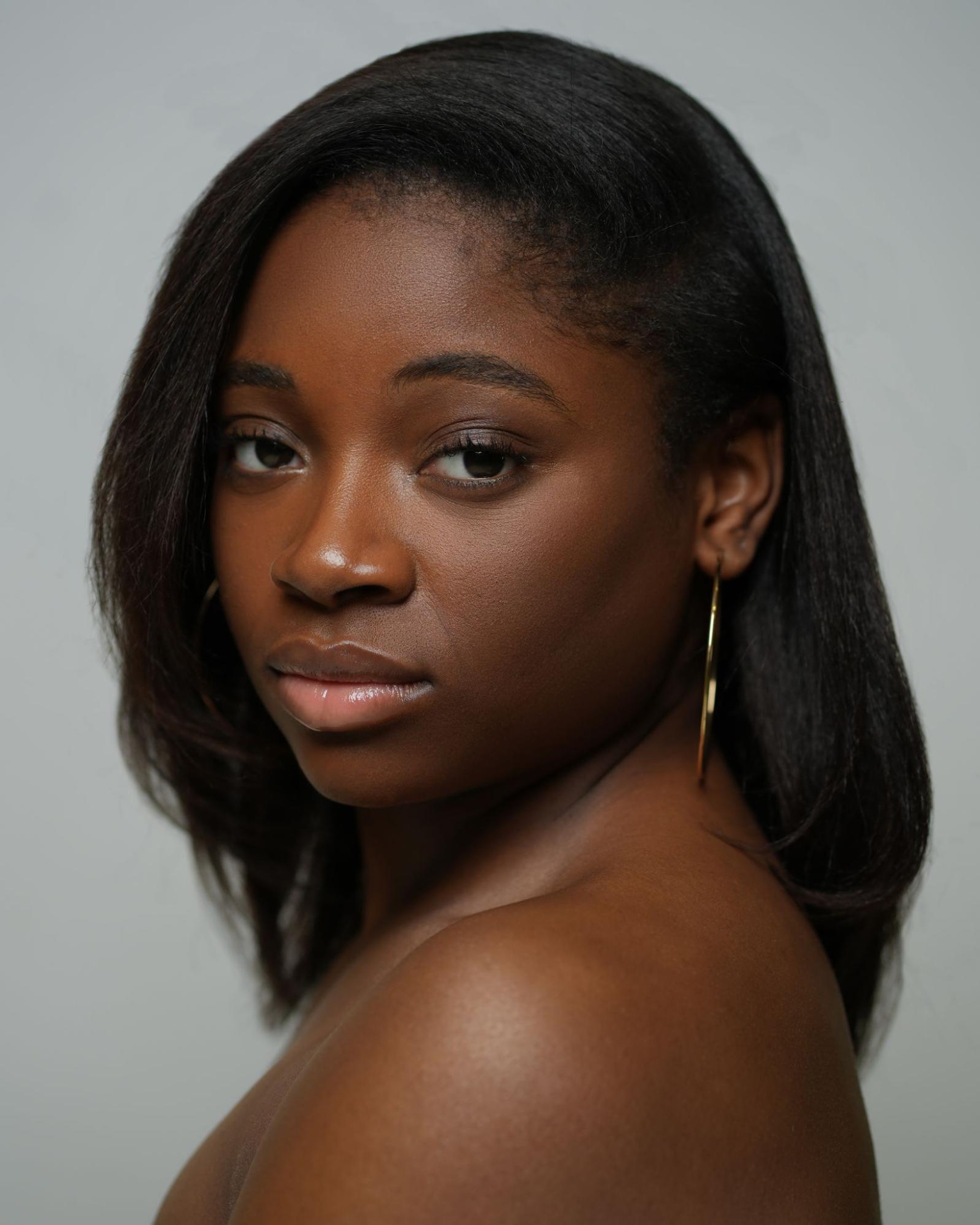 A portrait of dancer Spirit Priester, a Black woman who looks over one shoulder at us. She has straight hair down to her shoulders and looks at us with one eyebrow slightly raised. 