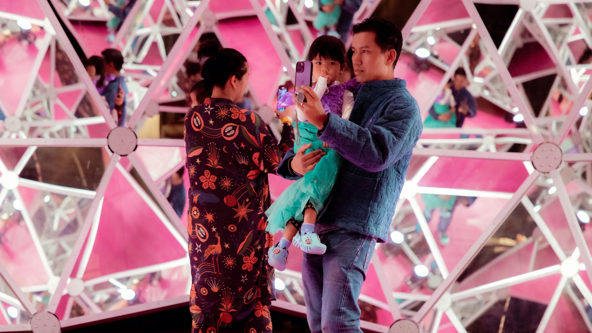 Visitors at Luna Luna stand inside a room covered by triangular mirrors, designed by Salvador Dalí. One of the couple holds a small child in his arms and holds up a phone to take a photo.