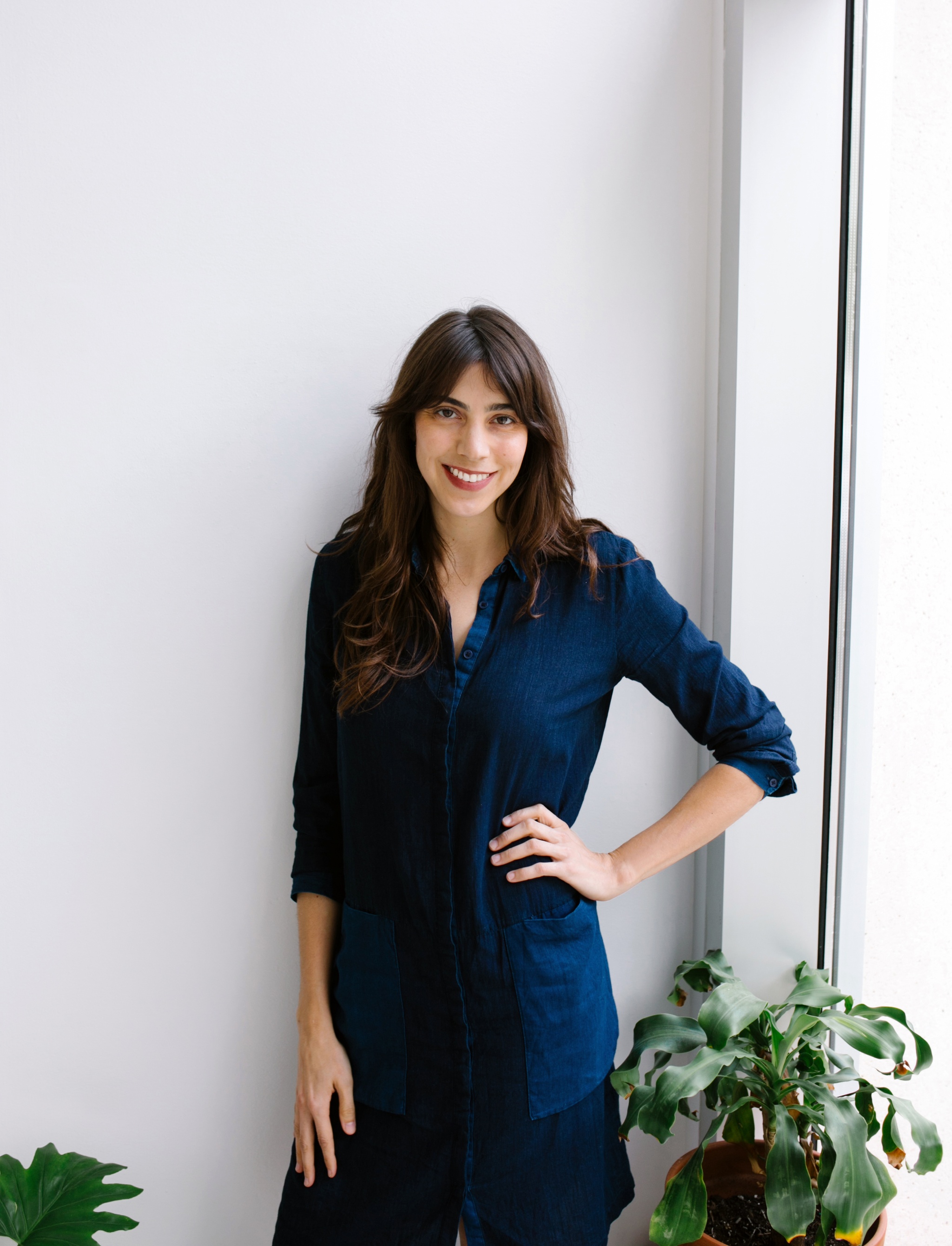 A photo of Arden Sherman standing beside a window with one hand on her hip. Green, leafy plants sit on either side of Arden. 