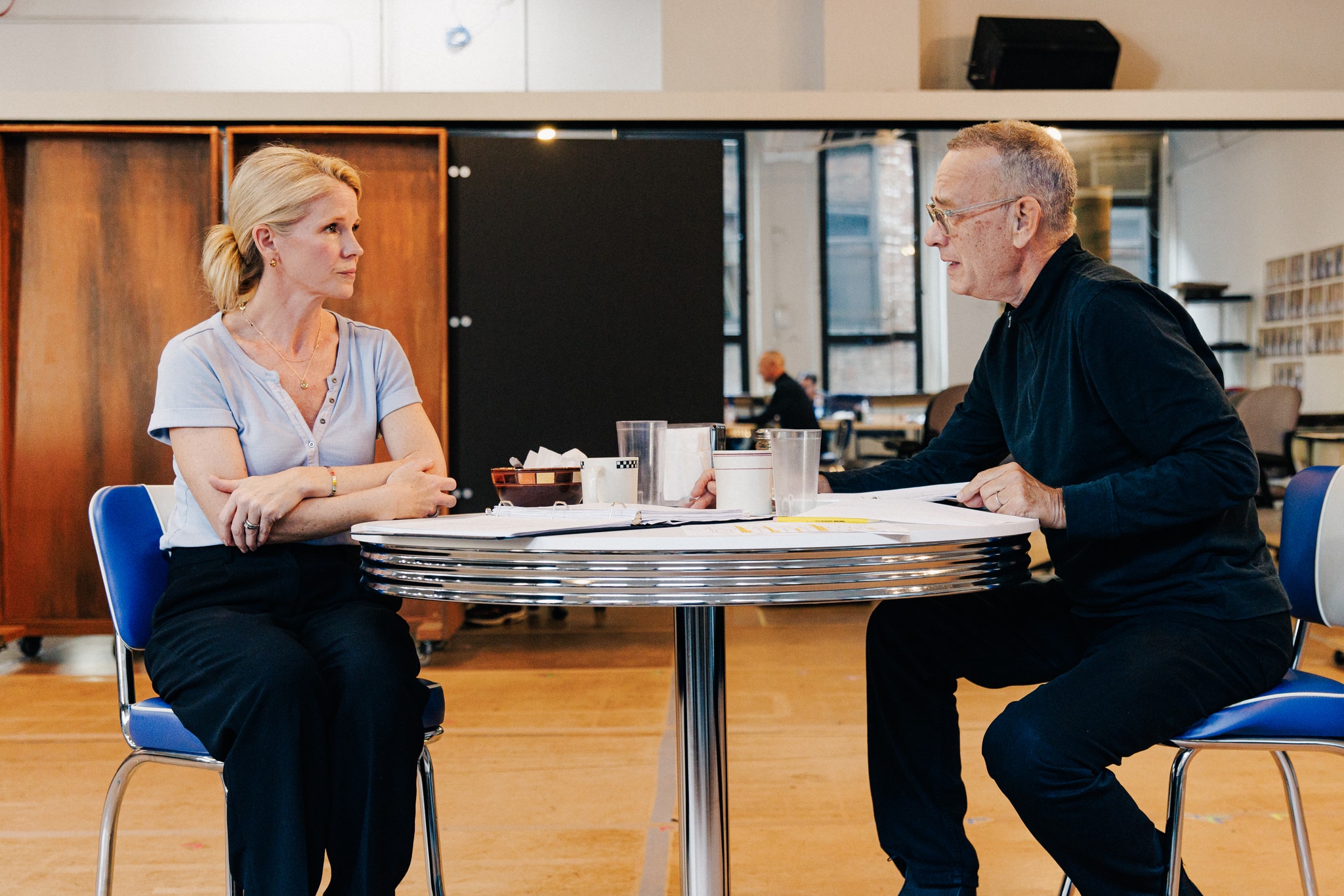 A photo of actors Kelli O'Hara, a blond white woman, and Tom Hanks sitting at a round diner table across from each other. They look intently at one another and we see them in profile.