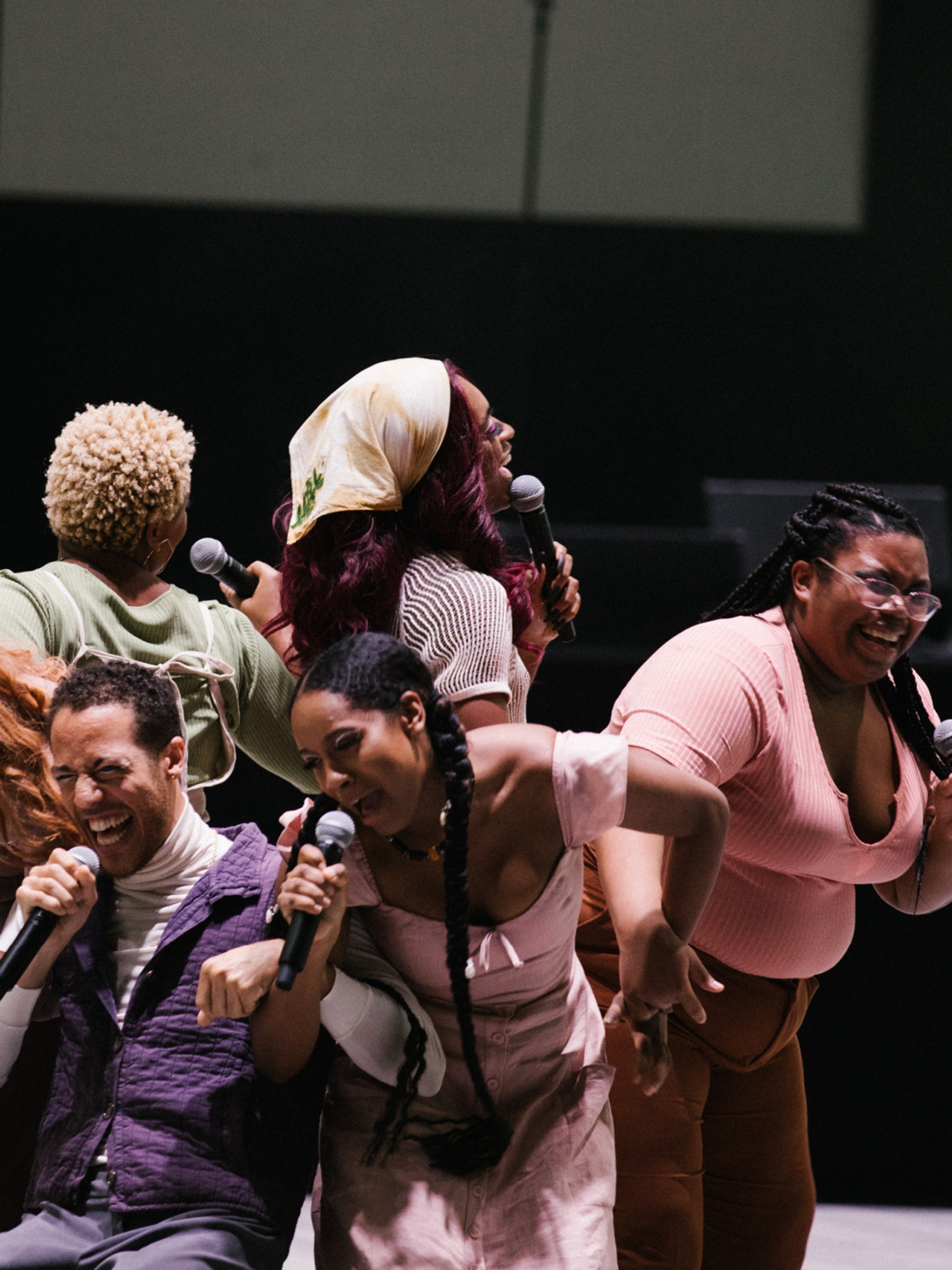 Five performers stand with their backs to each other in a circular formation. They all hold microphones and have strained looks on their faces as the speak into them. 