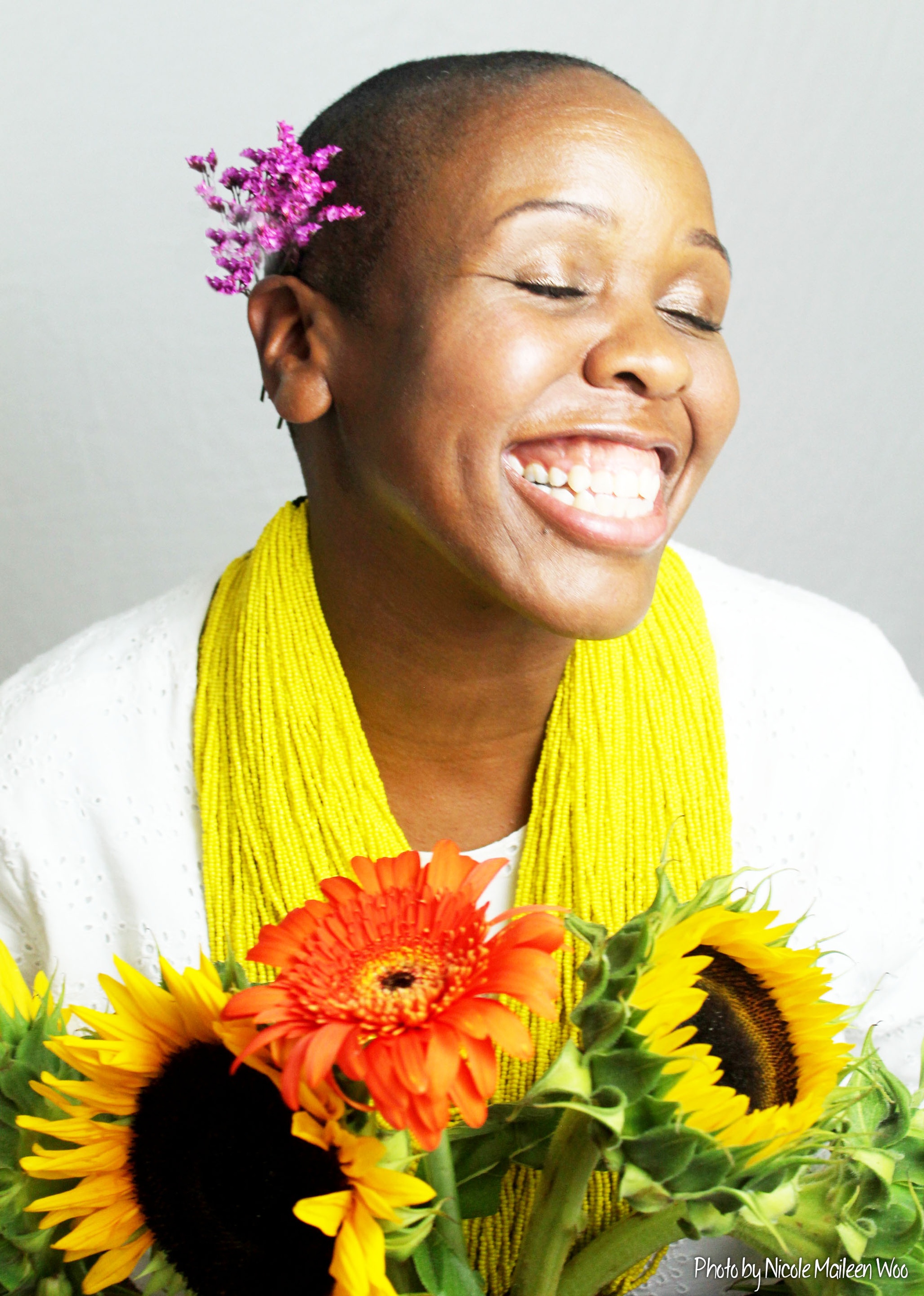 A portrait of artist Nehprii Amenii. Nehprii is a Black woman with a shaved head. She has her eyes closed and a wide smile as she turns her face joyfully toward a bright light. She holds a bouquet of sunflowers. 