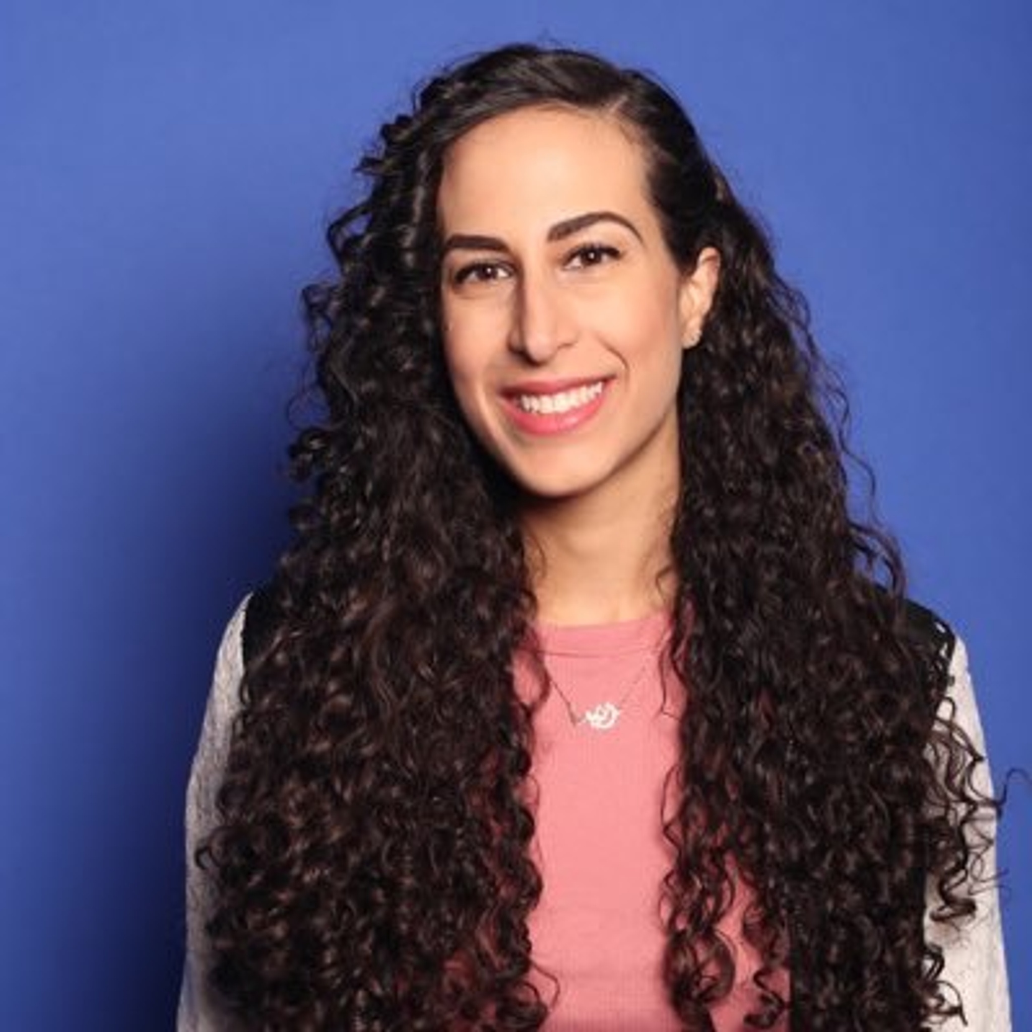 A woman with long curly hair that falls below her shoulders smiles against a blue wall. She wears a pink shirt and a necklace. 