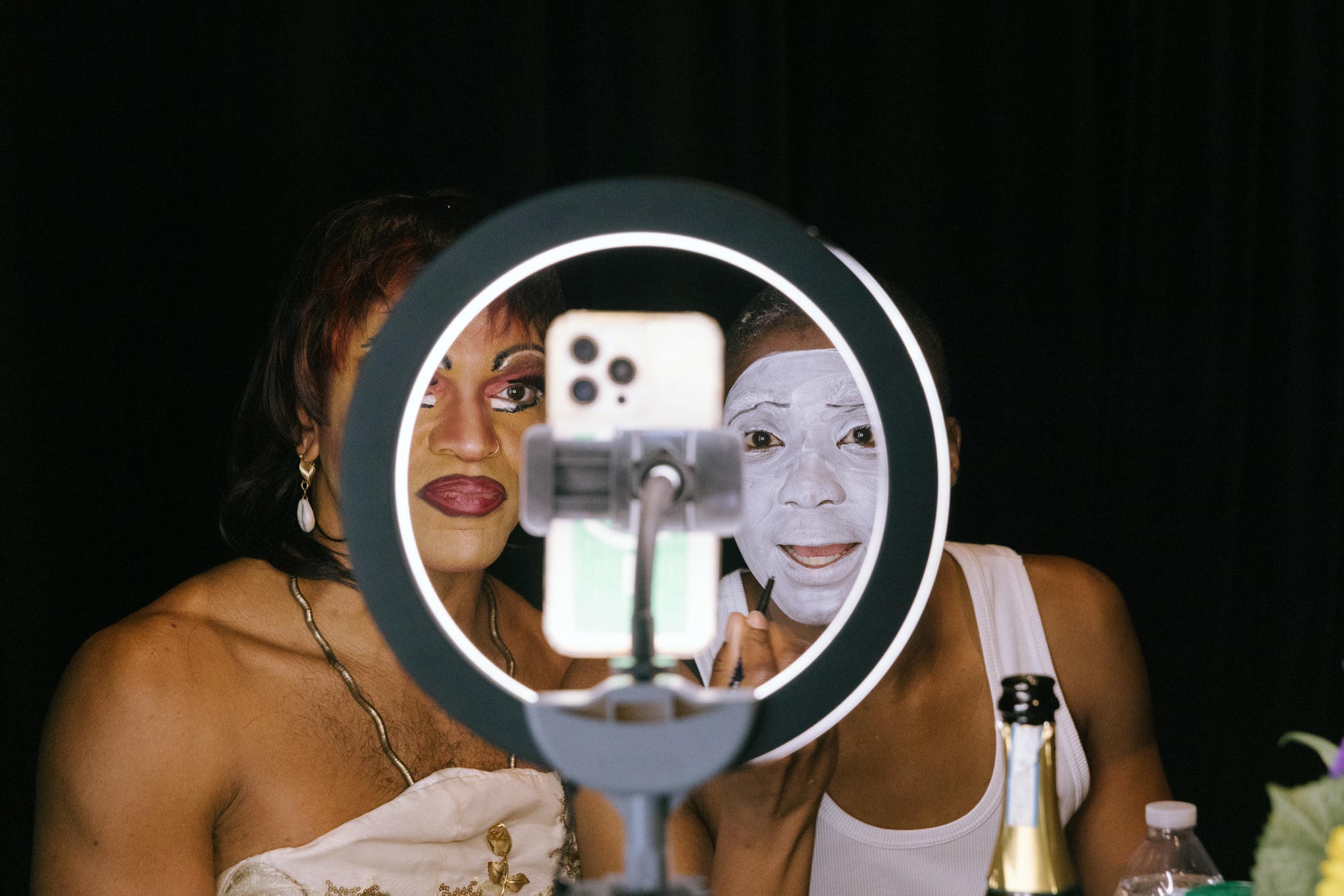The performers and artists Gabriele Christian and NIC Kay peer into an iphone held at the center of a ring light. We see them from the opposite side of the ring light, as if they are peering at us. Gabriele is in a short red wig, with a strong red lip and dramatic eye make up. NIC wears white face make up like a mime's and is applying eye liner.