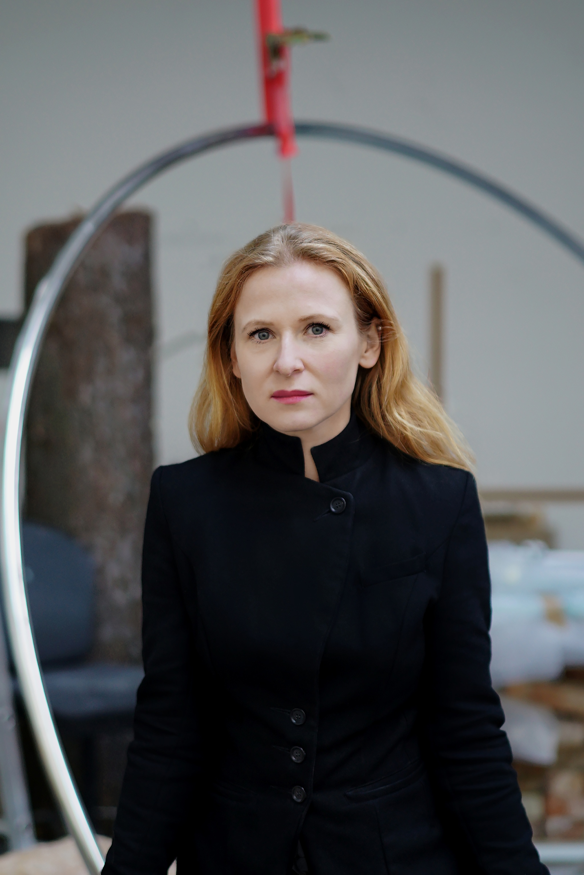 A white woman with blond hair flowing below her shoulders and behind her back looks into the camera through a thin metal hoop in the foreground of the image. She wears a high-collared black outfit. 