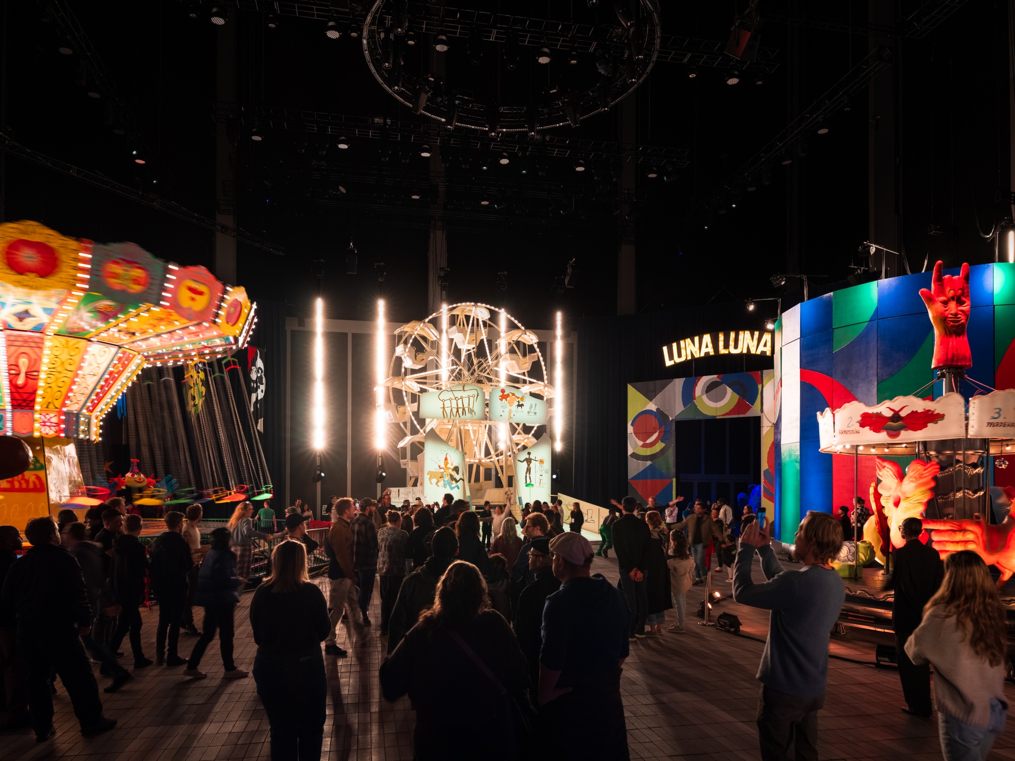 A group of gallery visitors are seen from behind as they move into a vast exhibition space containing art carnival rides and attractions. The rides include a colorful painted swing chair ride, a Ferris wheel, an entry archway, and a carousel.