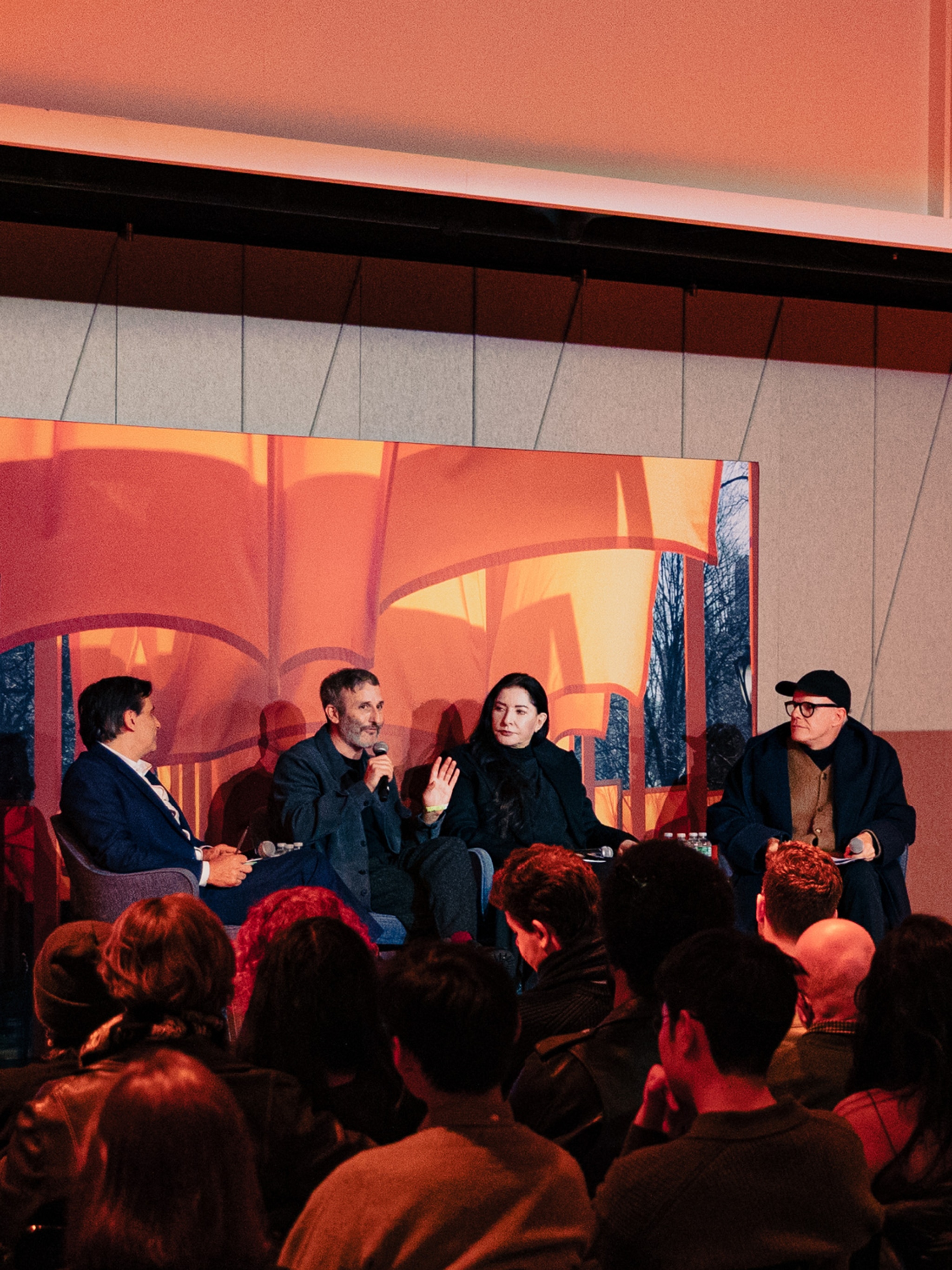 Four conversation panelists sit on a stage with a background behind them showing the saffron colored fabric of Christo and Jeanne-Claude's The Gates installation.
