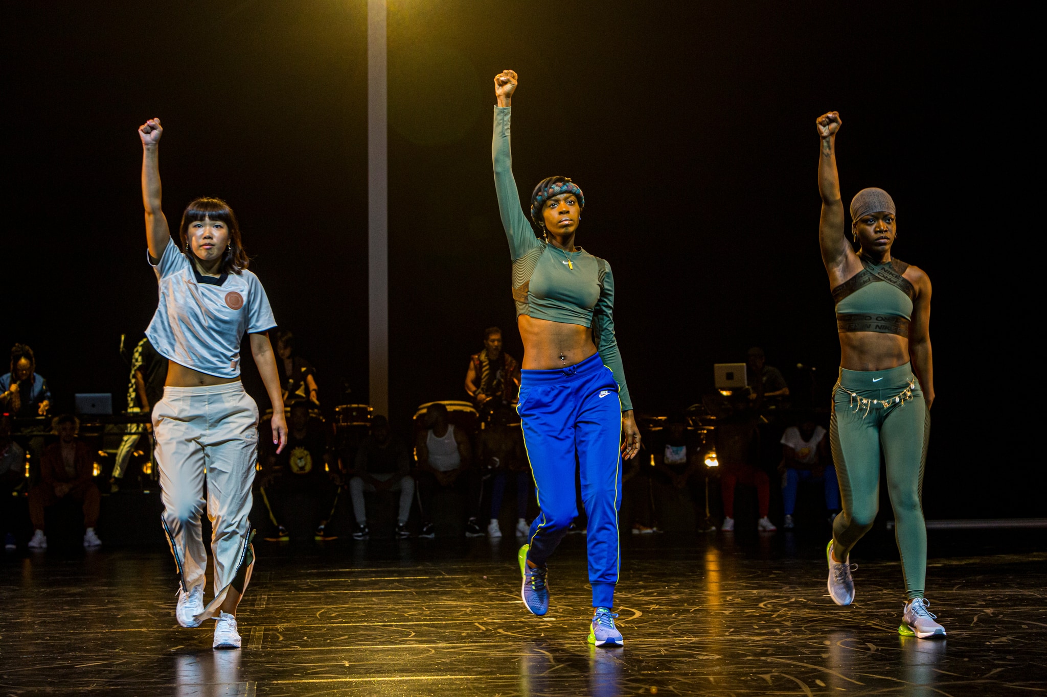 Three women dancers stand in a row on a dark stage. They are spaced with about 3 feet between them and each stand straight and tall with their right fists raised. 