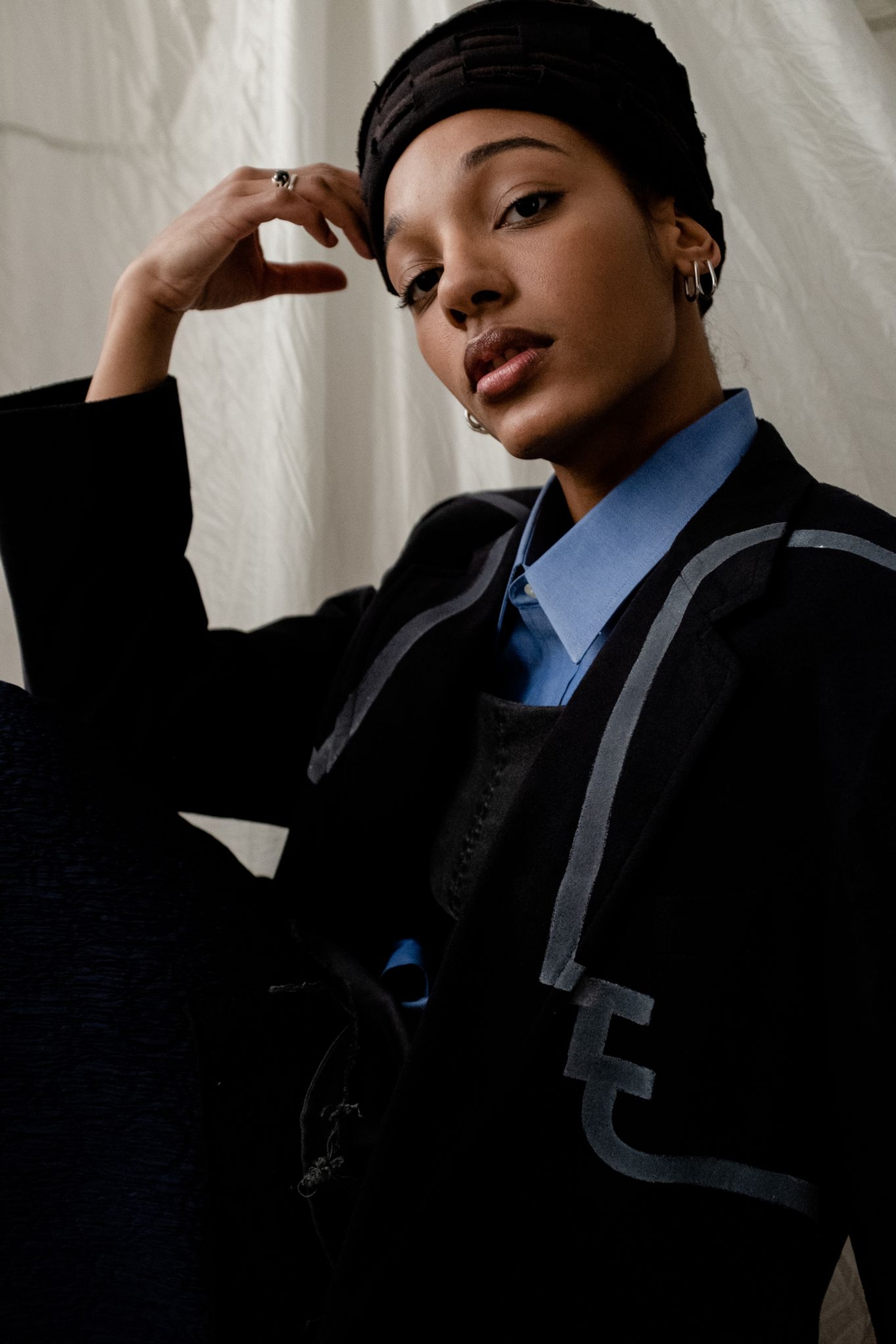 A portrait of Afro-Dominican dancer and choreographer Katherine Paola De La Cruz. Katherine poses in a seated position with one leg drawn to her chest. She rests her elbow on her knee and leans her head one one hand. She wears a jacket over a blue button down blouse. She looks directly at us with eyebrows slightly raised.  