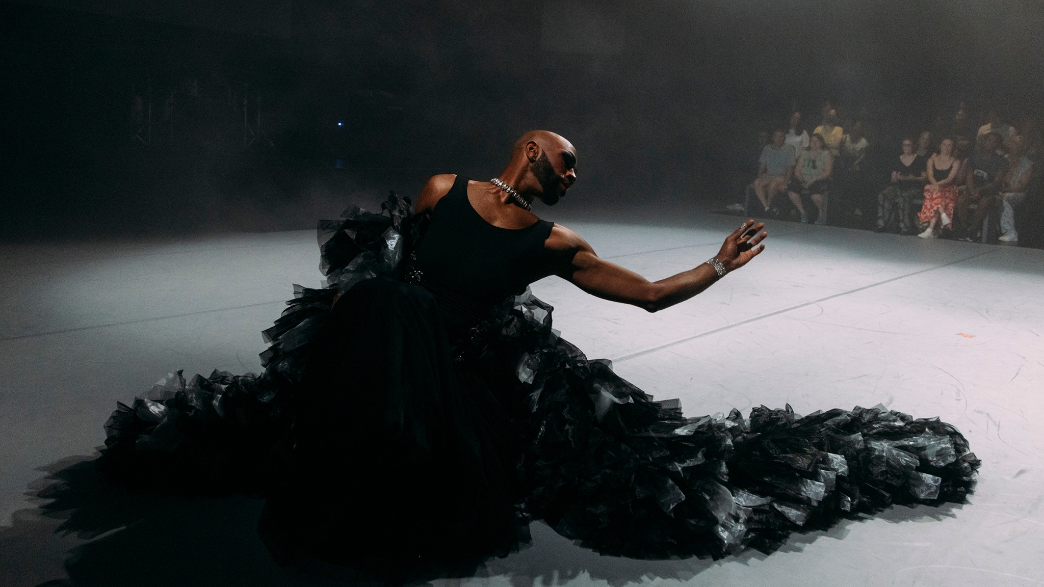 Cain Coleman, a Black performer and artist, dances on stage during the performance of "New Information." Cain wears a ruffly, billowing black gown, and seems to sink into it on the floor, one arm gracefully outstretched. 