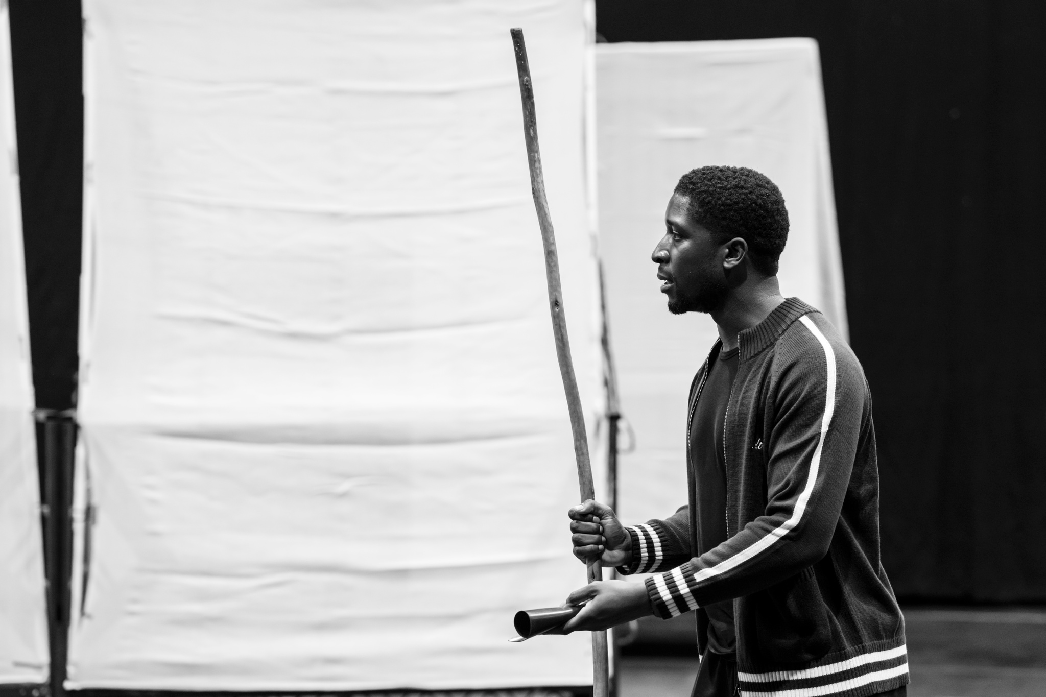 Actor Caleb Obediah, a Black man, in rehearsal. He wears a track jacket and holds a tall prop staff. He is seen in profile holding one hand open out in front of him. 