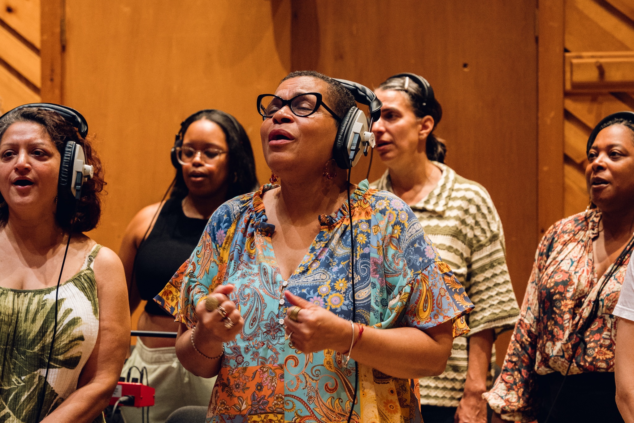 A member of the choir, a Black woman wearing a flowy, blue-and-gold floral printed shirt, sings into a microphone. Her face is slightly upturned, and her eyes are closed in concentration. She holds her hands up in front of her chest, embodying the energy of her song.