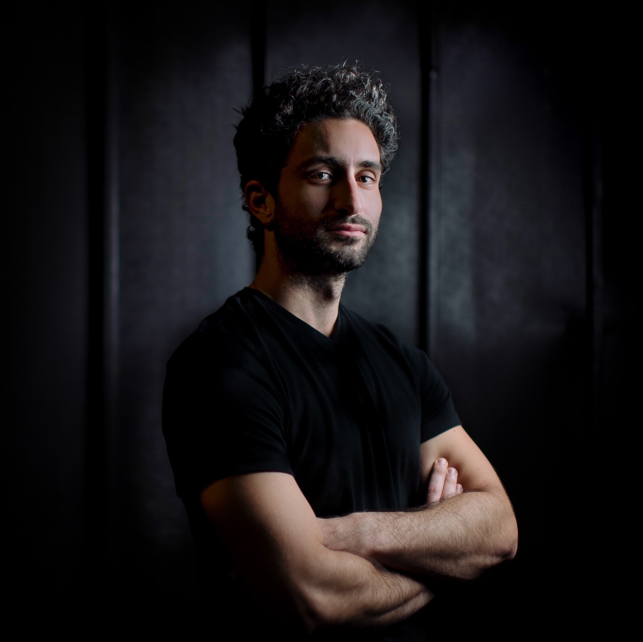 A portrait of Turkish-American artist Avi Amon. Avi poses against a dark gray background. He wears a black t-shirt and crosses his arms over his chest. He has curly salt-and-pepper black hair and turns his head slightly to look at us directly with a mysterious look on his face. 