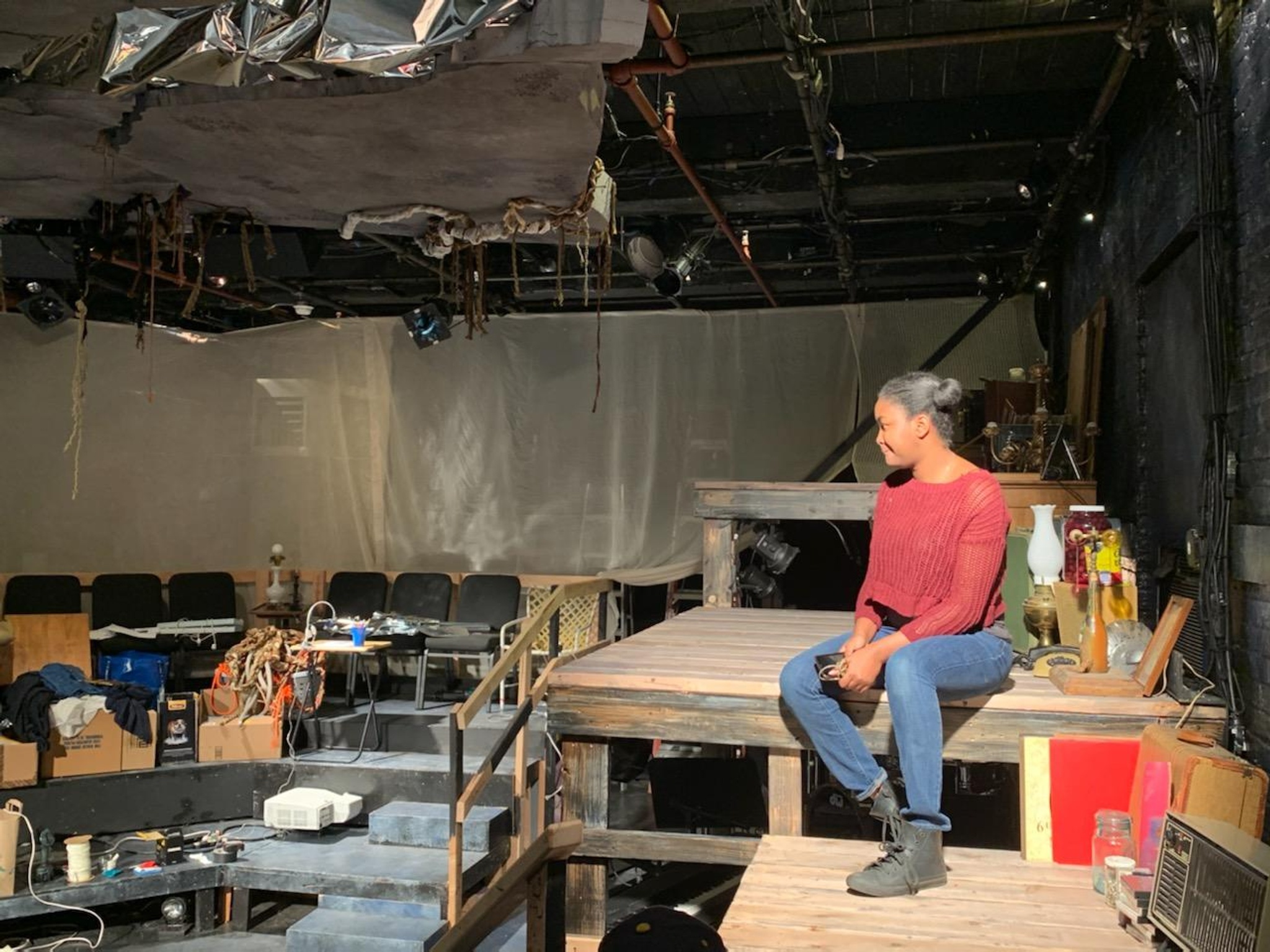 A woman in a red sweater and blue jeans sits illuminated in a stagelike structure in a black box theater that appears to be under construction