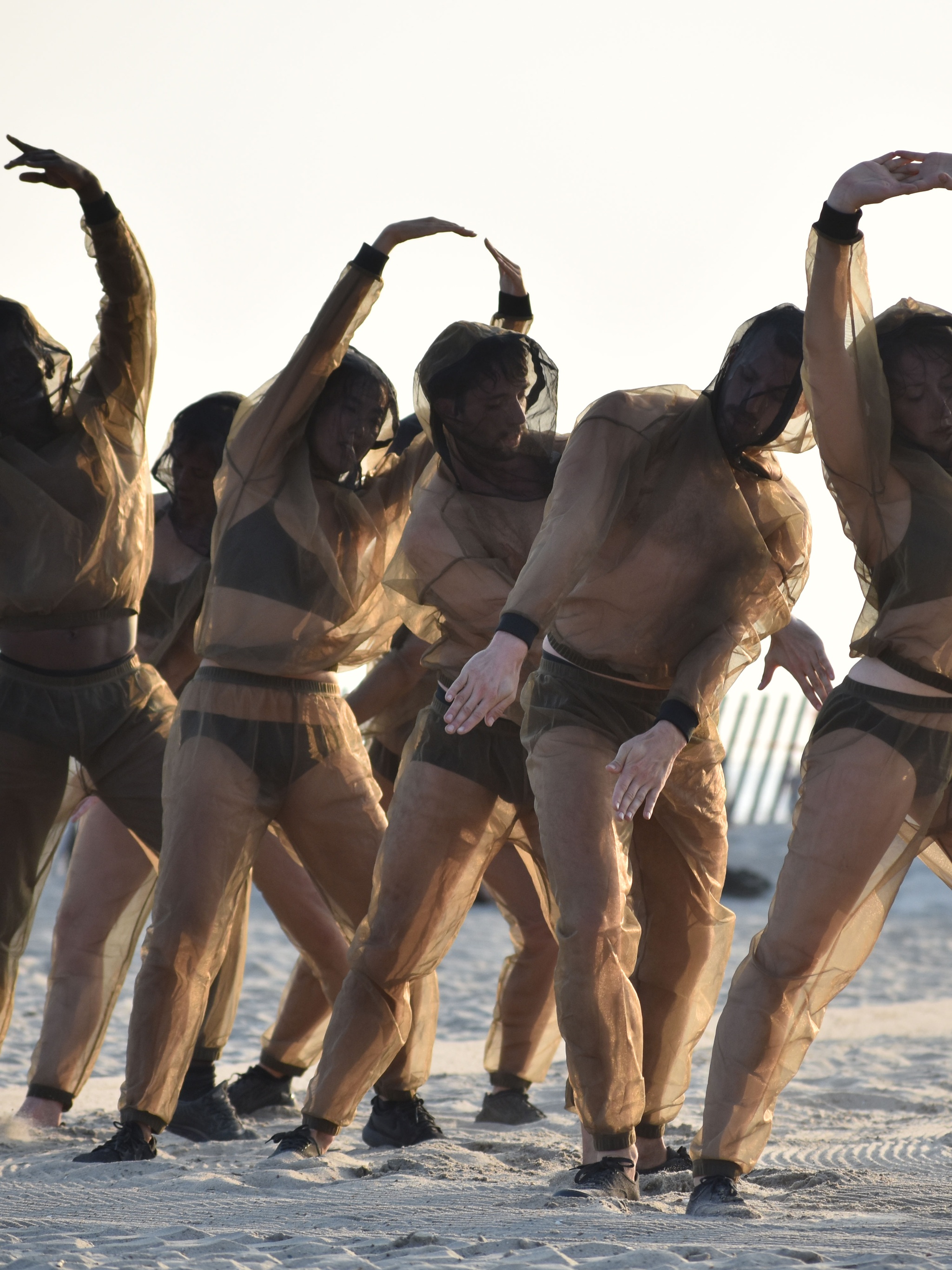 Dancers in sheer brown outfits dancing outdoors on sand.