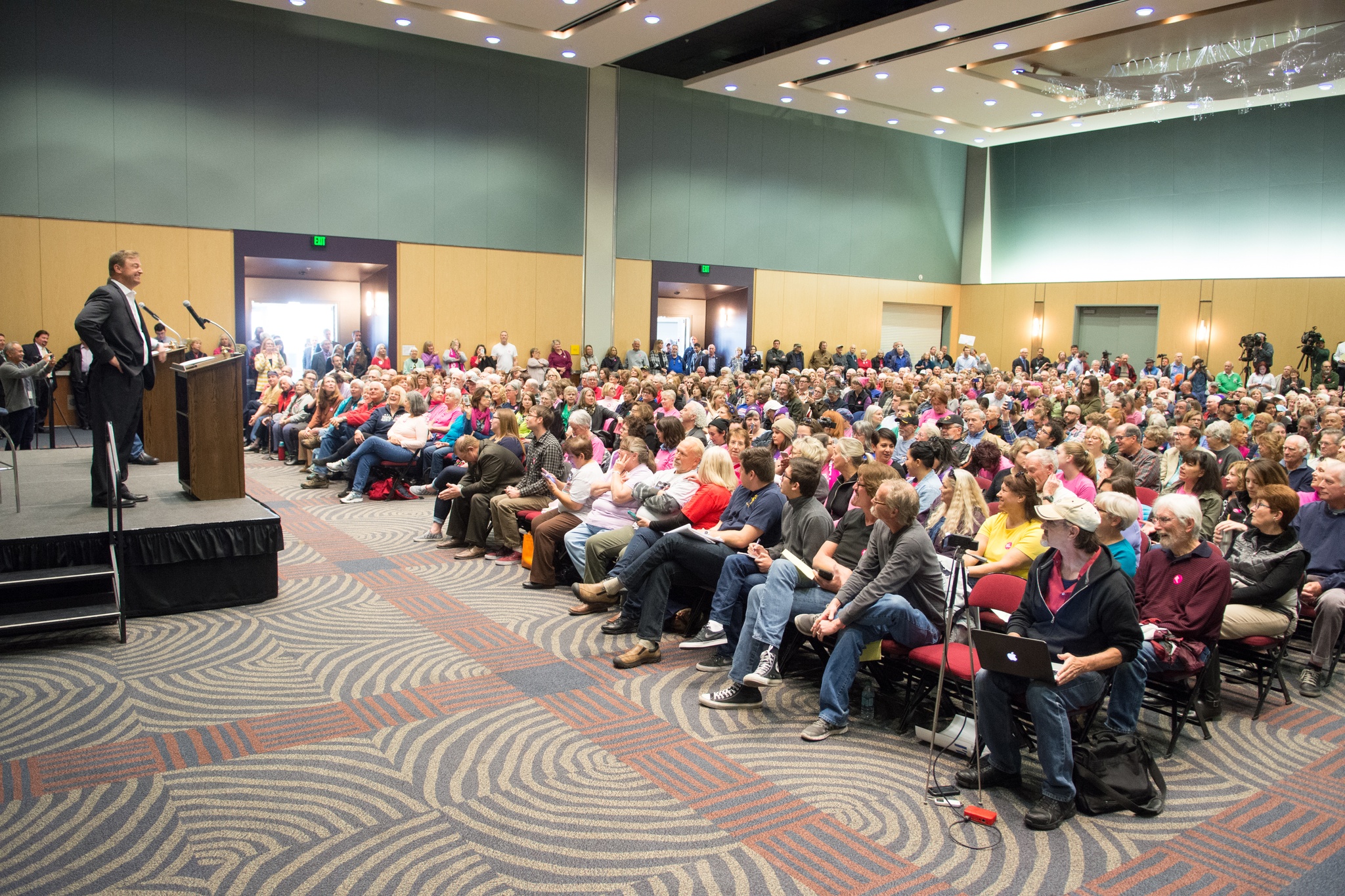 An audience in a hall listens to a man at a lectern on a stage in front of them