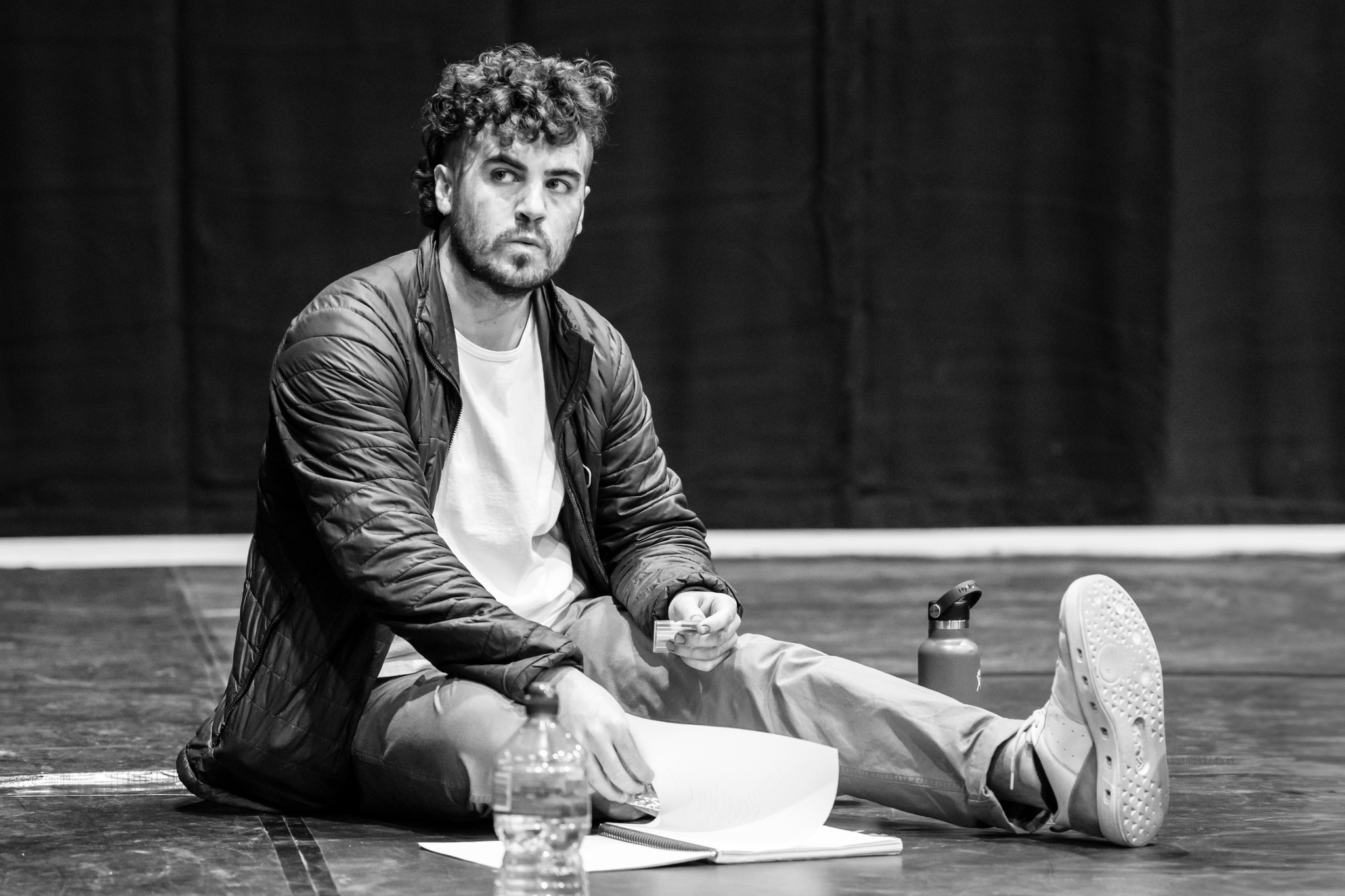 Actor Ian Bouillion sits on the floor of a rehearsal room. He has curly dark brown hair that falls over his forehead. He sits casually with one leg tucked in and one stretched out in front of him. 