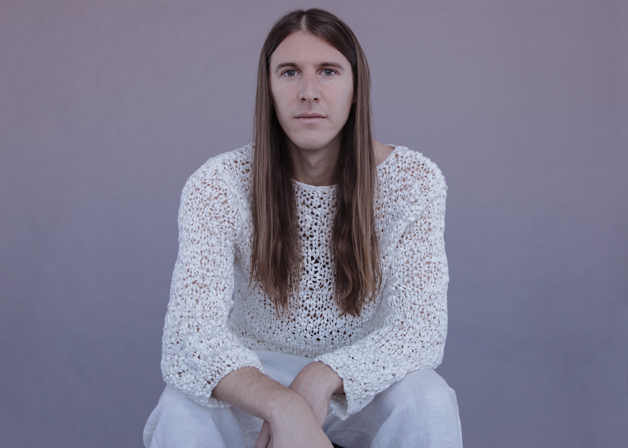 A person with long brown hair hanging over their shoulders, sitting with elbows on their knees and hands crossed between them. They wear a see through white knit top and white pants.