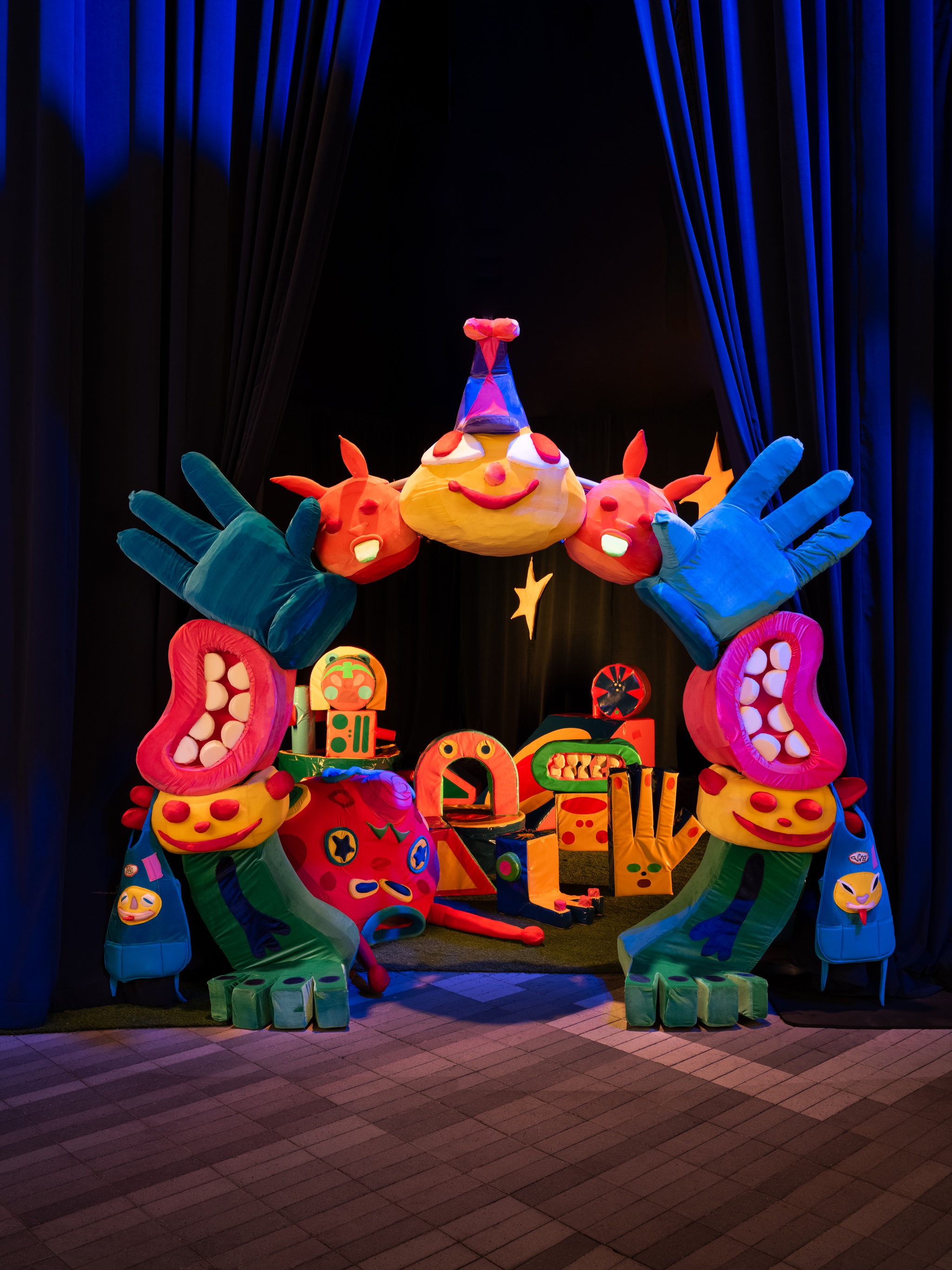 A colorful entryway opens in a gallery exhibition hall in PonciliCreación's installation. The entry is decorated with cartoonish smiling faces, open hands, and mouths with pink lips and white teeth. Inside are large foam objects in similar shapes and colors.