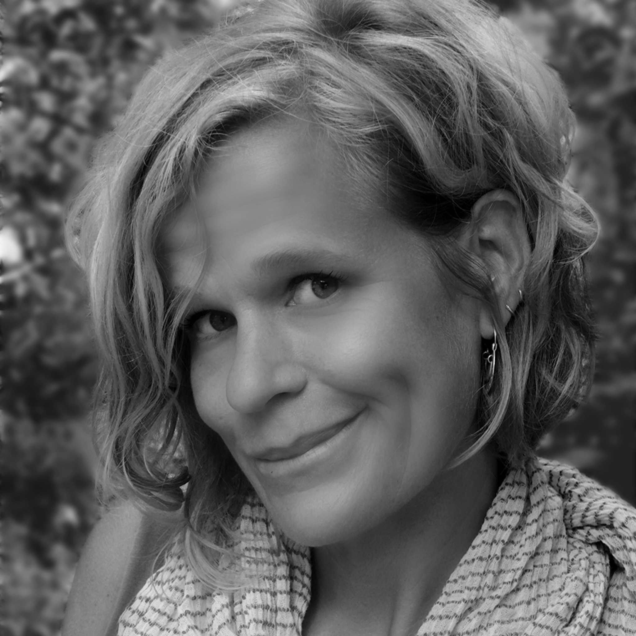 A black-and-white photo of Betsy MacLean. In the tight close-up, MacLean grins at the camera in a three-quarter view of her face from the left. Her hair is short and wavy, she wears a scarf loosely around her neck, and blurry foliage is just discernible in the background.  