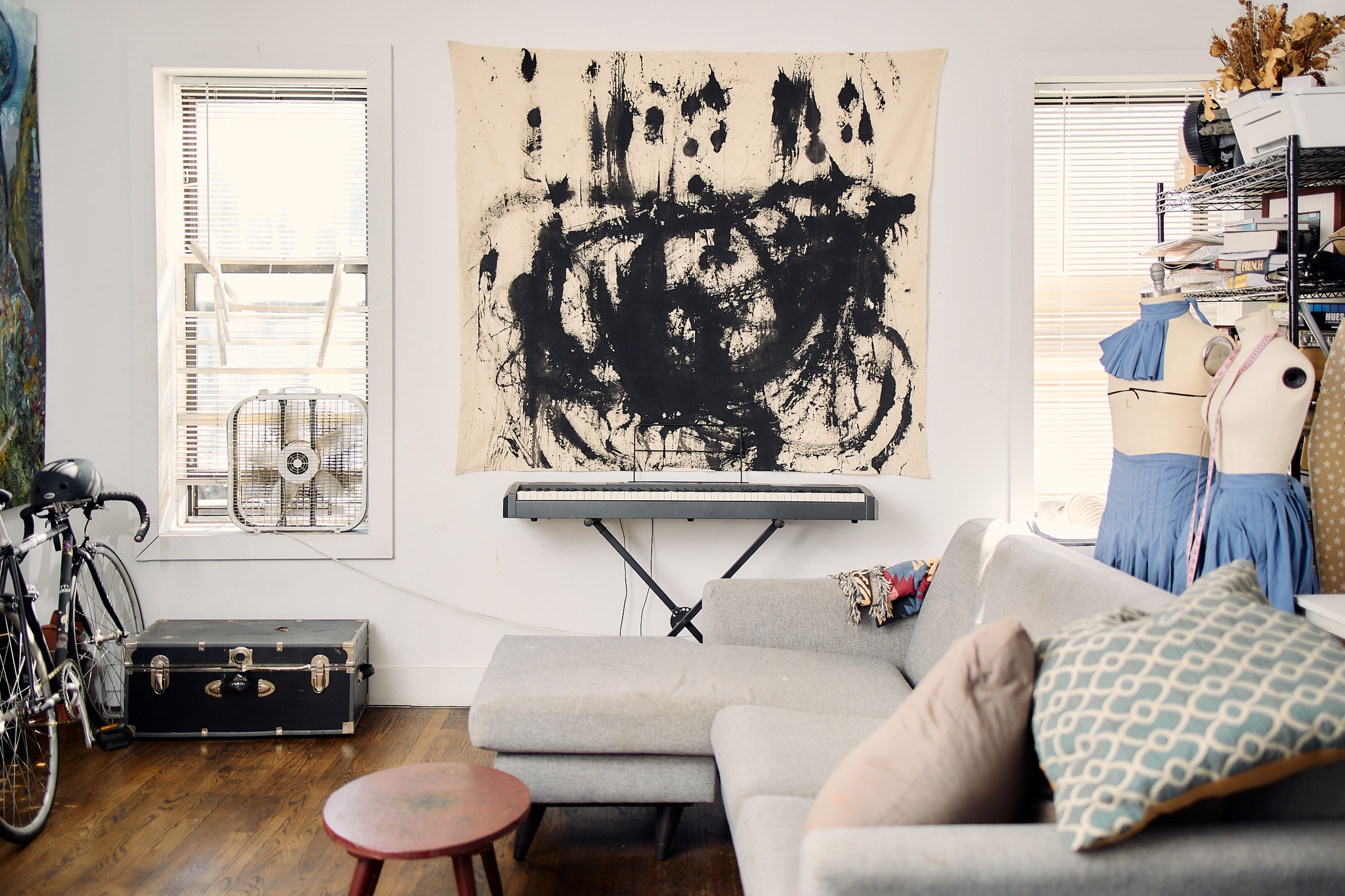 An abstract painting by Jarrett Key with dramatic swirling gestures of black paint hangs above a piano keyboard and a long sectional sofa in the artist's studio space. The space is filled with light from two windows on either side of the painting. 
