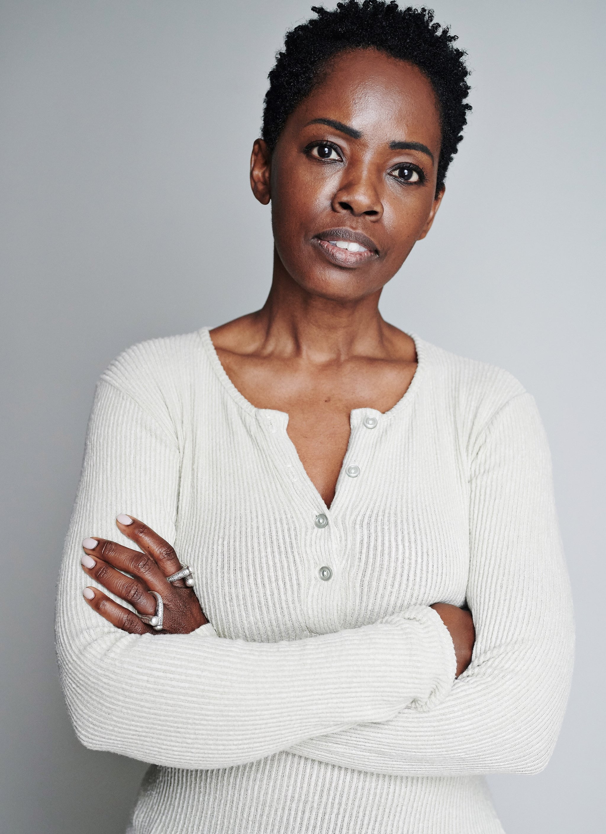 A photo portrait of actor Lorna Brown. Lorna is a Black woman with short dark hair. She wears a long sleeve white shirt, and looks directly at us with mouth slightly open and eyebrows slightly raised. She crosses her arms as she tilts her head slightly to the right. 