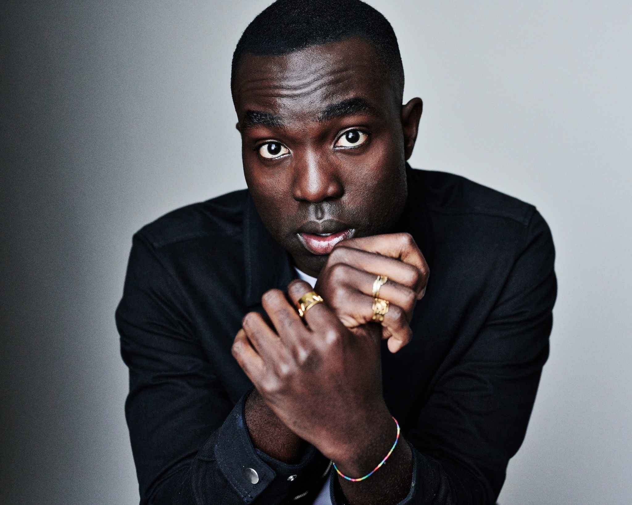 A portrait of actor Paapa Essiedu seen from the chest up facing us. Paapa, a Black man with short hair, sits with hands raised in front of him, shoulders slightly slumped. He looks directly at us with eyebrows raised. He wears a deep navy blue jacket. 