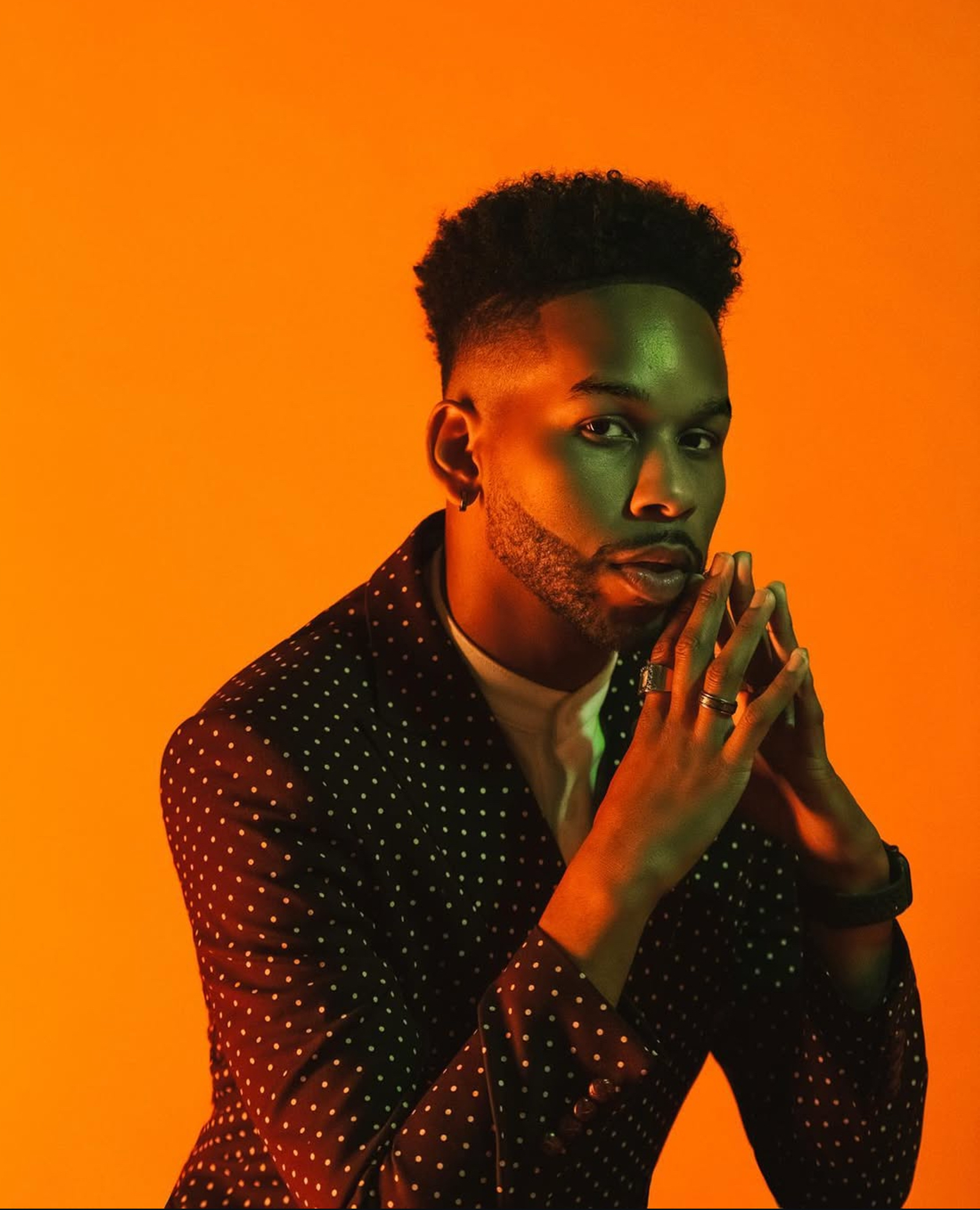 A portrait of actor Israel Erron Ford, who poses against a deep orange backdrop. Israel is a Black man whose body is bathed in a glowing orange light as he poses in a three-quarter pose, peering at us from his left, with hands raised together to his chin. He wears a polka dot patterned suit jacket and wears his hear in a hi top fade. 