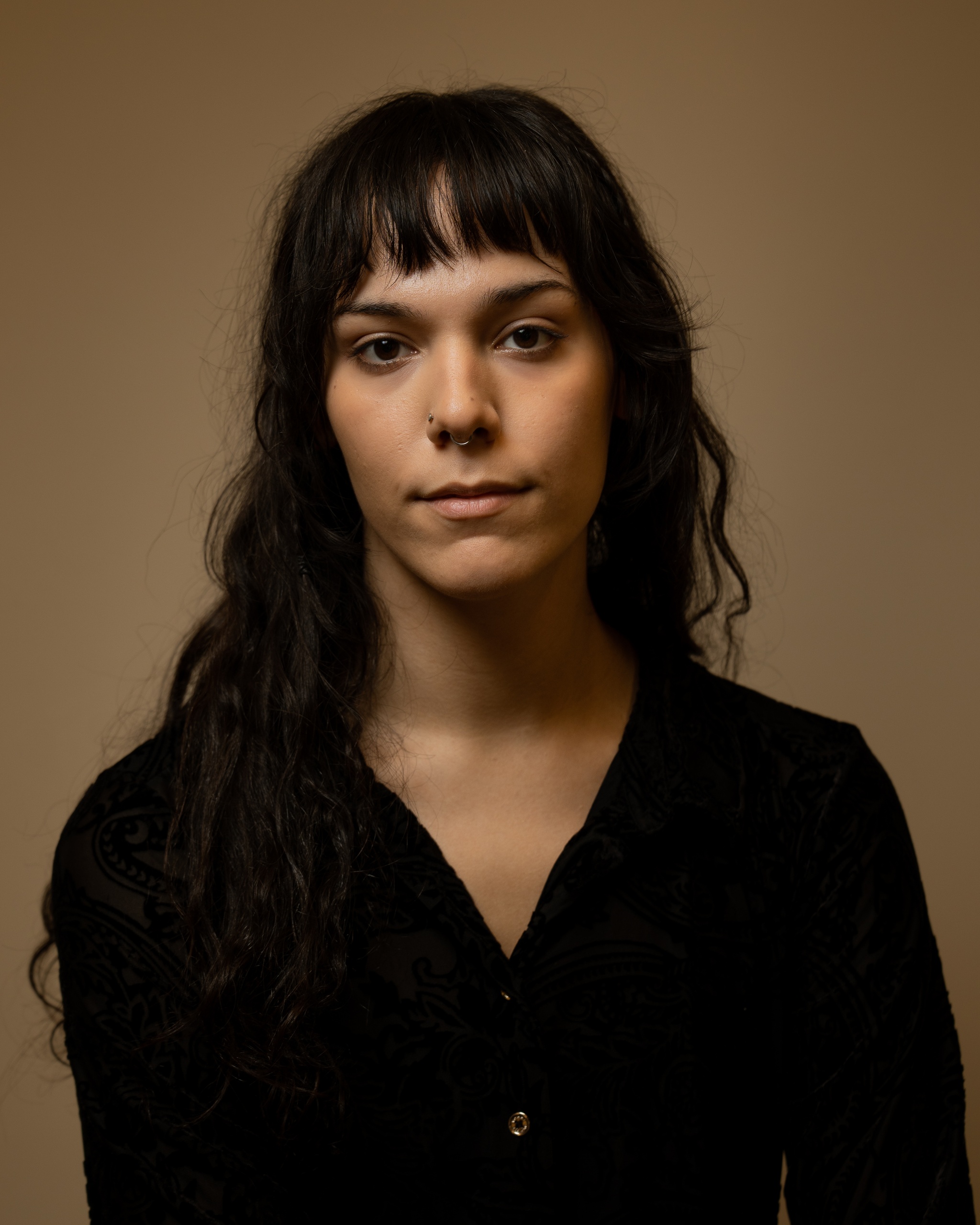 A portrait of Dahlia Khair against a neutral brown background. Dahlia has dark brown hair that is wavy with bangs across her forehead. She looks directly at us with a slight smile at the corner of her mouth.