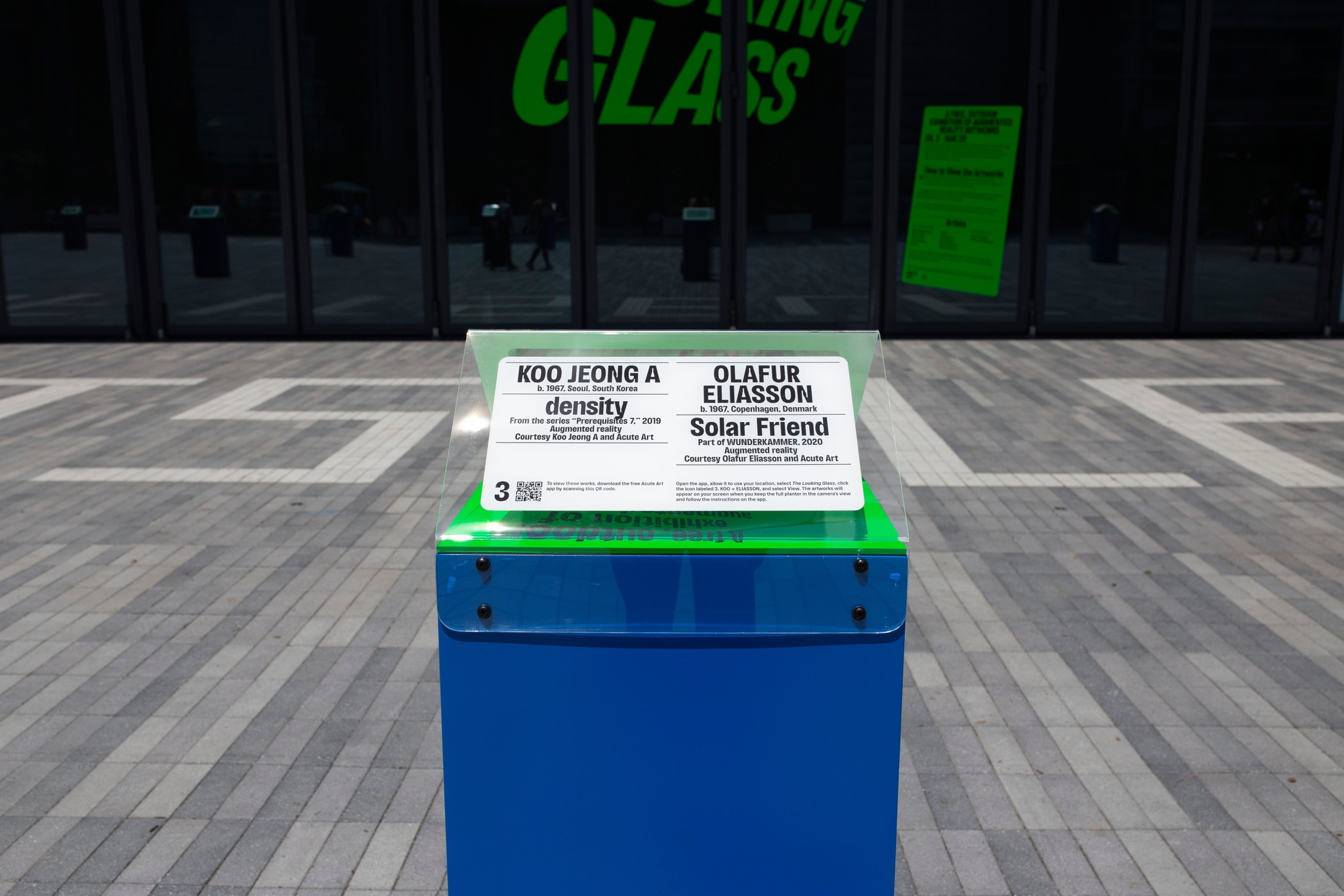 A bright blue plinth with a triangular top with an art object label on an outdoor plaza