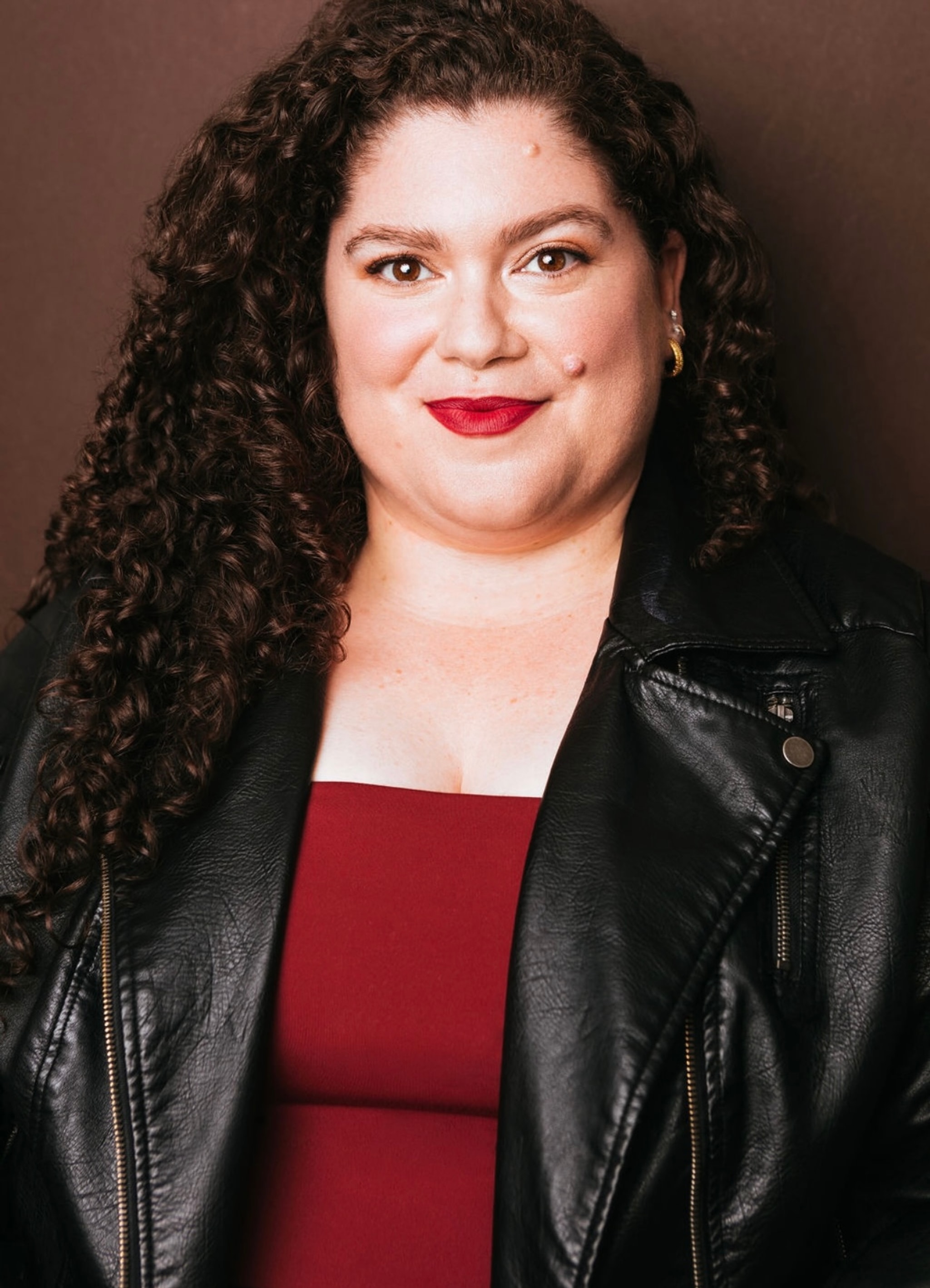 A portrait of Rebecca Spigelman, a white actor, who is seen from the waist up against a neutral backdrop. She has long curly dark brown hair. She wears a deep red tube top under a black leather jacket and her lip color matches the tube top. She smiles, gazing directly at us.
