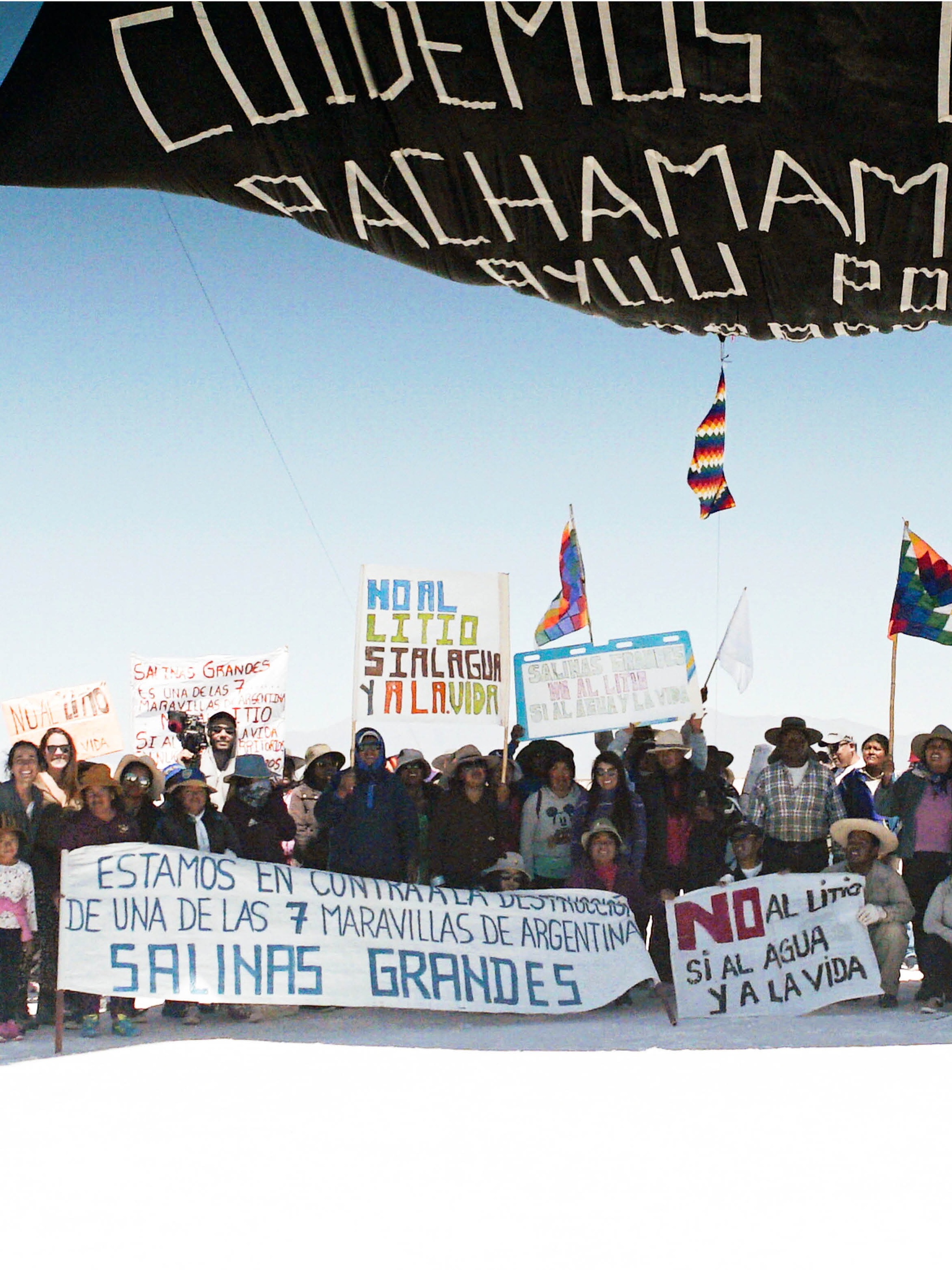 A group of people gather with activist banners beneath a black hot air balloon on a white salt flat