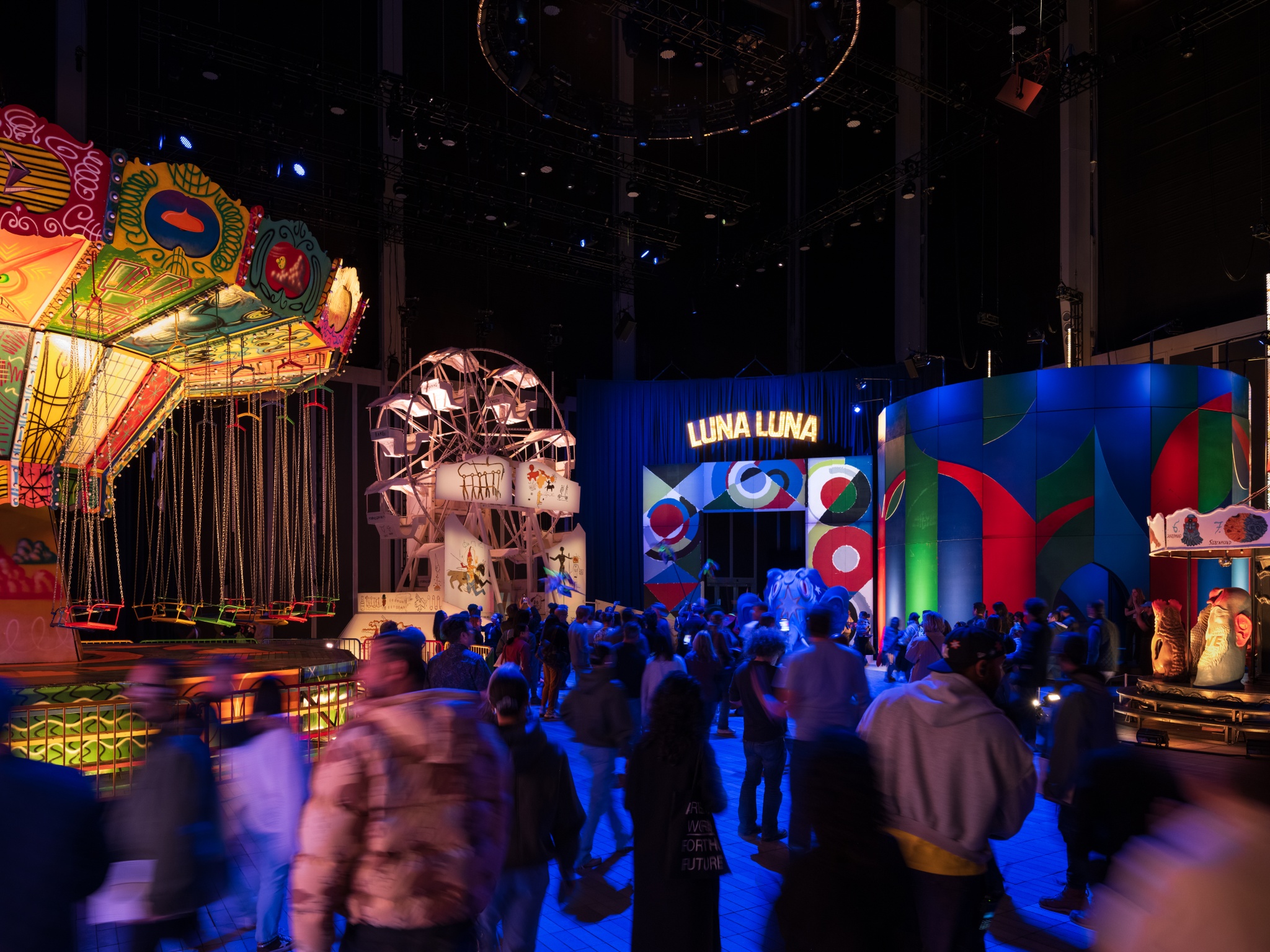 A group of gallery visitors are seen from behind as they move into a vast exhibition space containing art carnival rides and attractions. The rides include a colorful painted swing chair ride, a Ferris wheel, an entry archway, and a carousel.