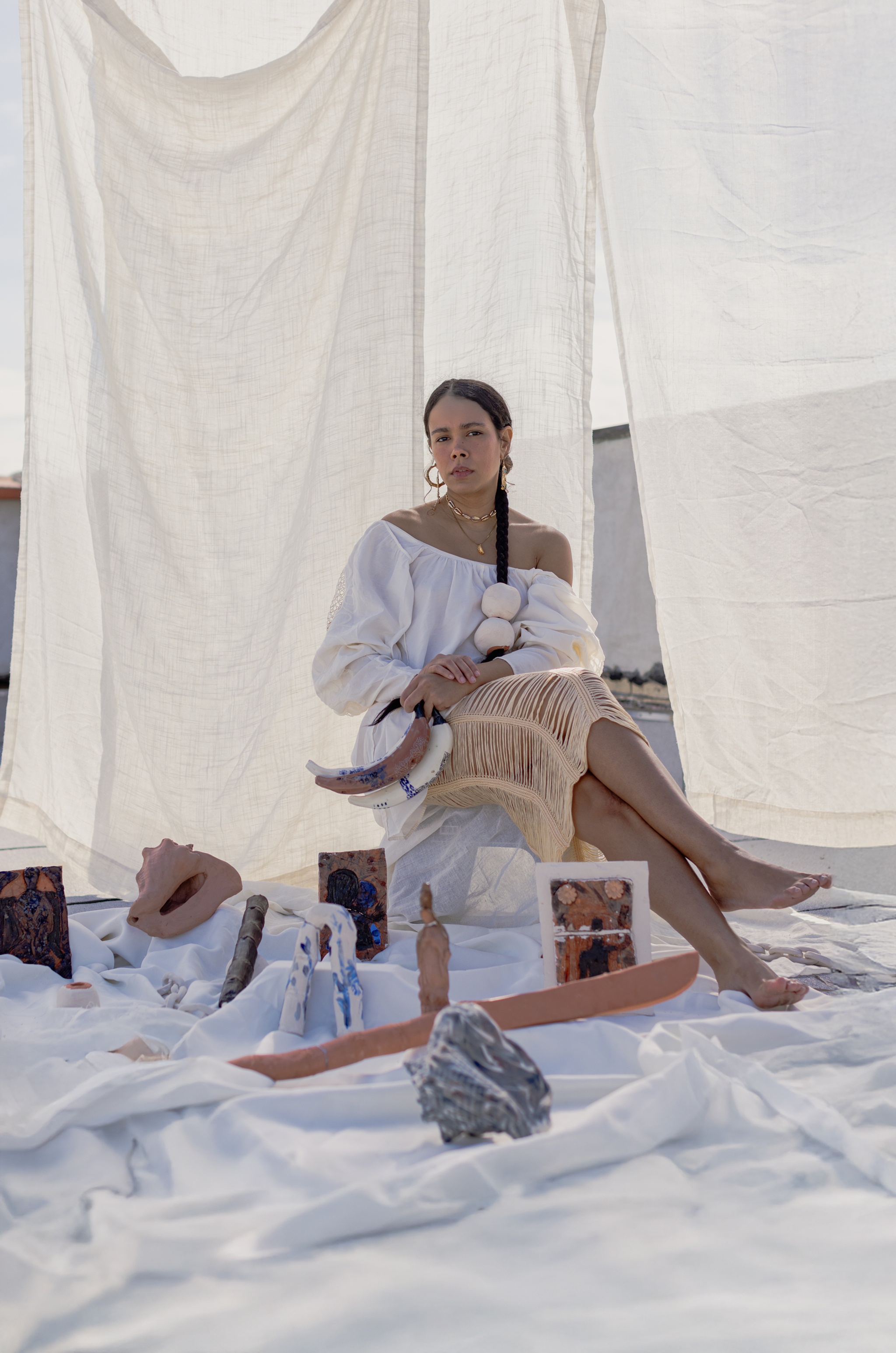 A portrait of Afro-Dominican artist Patricia Encarnación. Patricia is dressed in white loose clothing, with a shoulderless blouse. She sits with legs crossed in a space covered in white fabric and white sheer curtains. Around her feet are art objects including small paintings and a sculpted conch shell. 