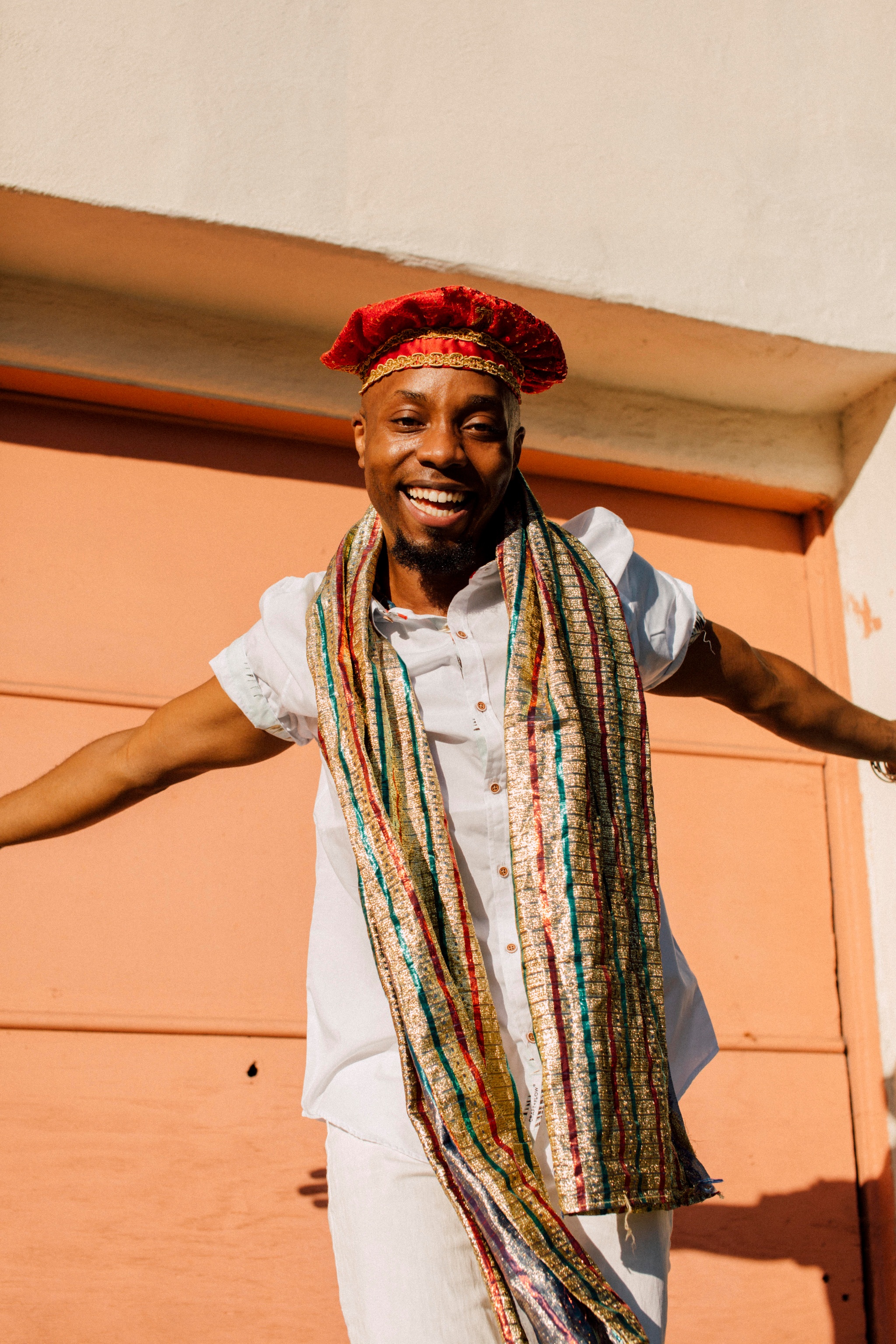 A Black man wearing a floppy red hat with arms outspread wearing a long multicolored scarf around his neck. He is in full sunlight and his shadow appears on the wall behind him. 