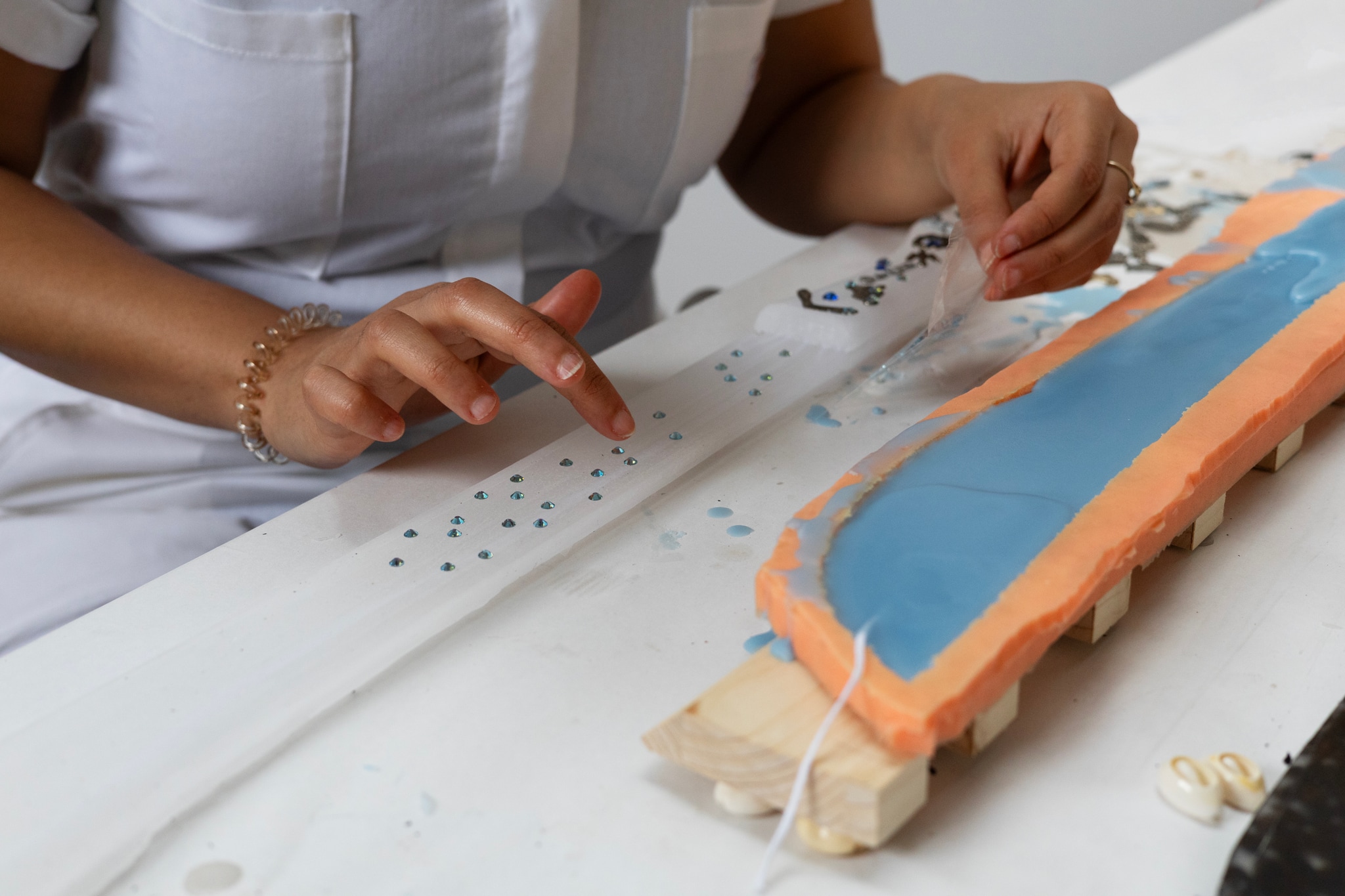 The hands of artist Yelaine Rodriguez are seen delicately attaching blue sequins to a strip of gauzy white fabric. The fabric is stretched out over a table that Yelaine, who we see only at waist level with her hands at work, leans over. 