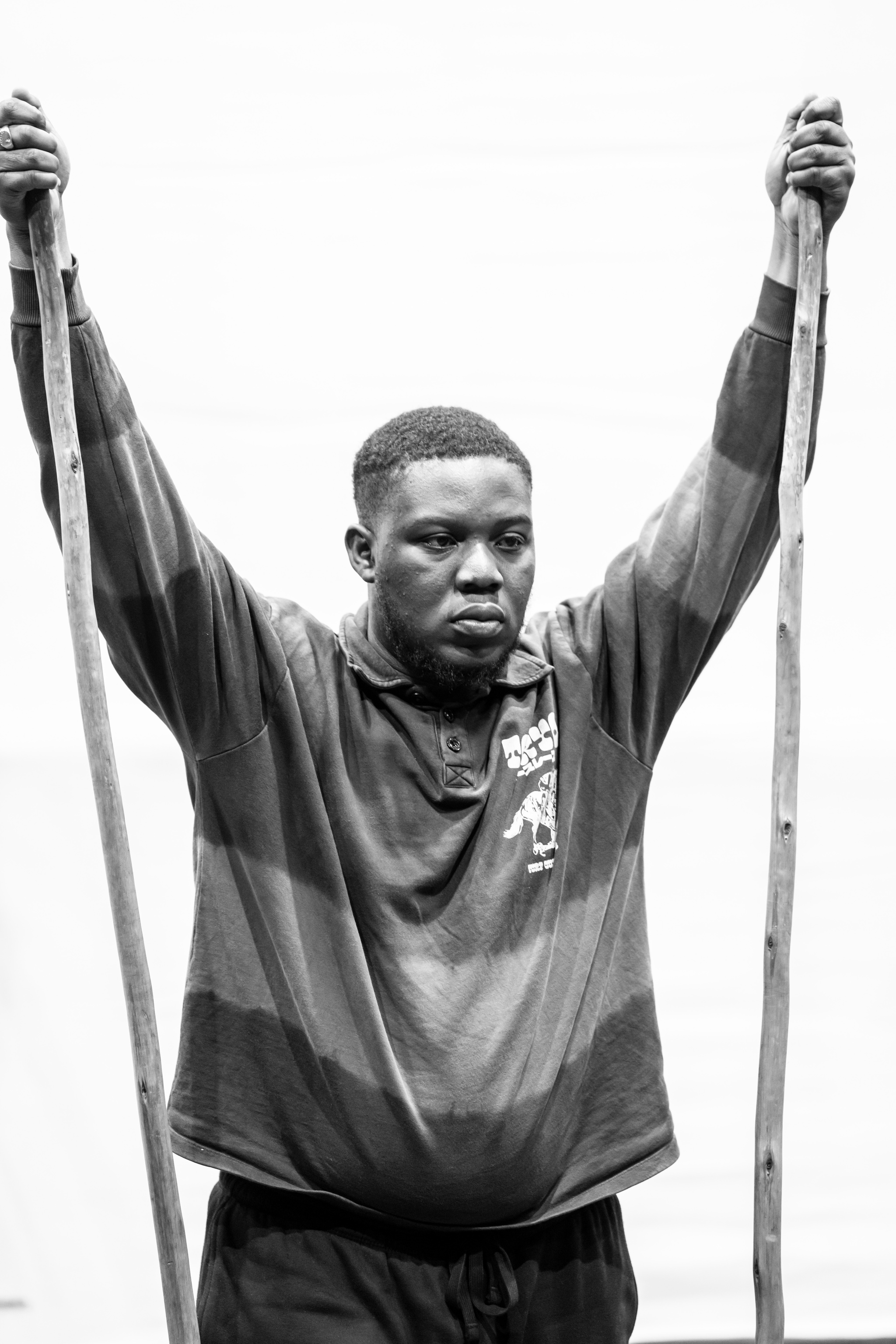 Actor Raymond Anum, a Black man, stands in his own clothes in rehearsal holding two tall staffs. His arms are raised up holding a staff on either side of himself. 