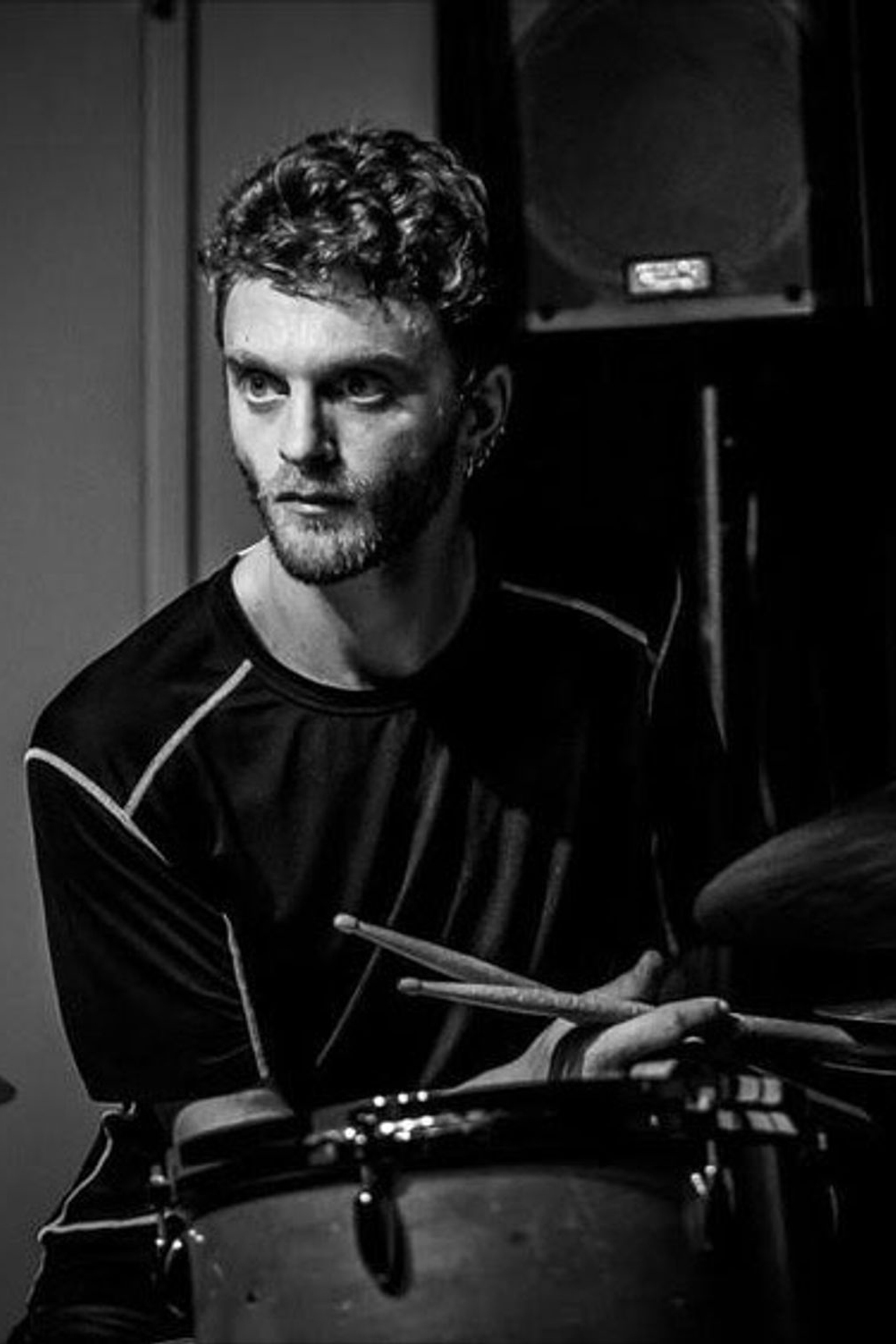 A white man with short curly hair sits behind a drum set in a black and white photo. He looks intently away to his right side.