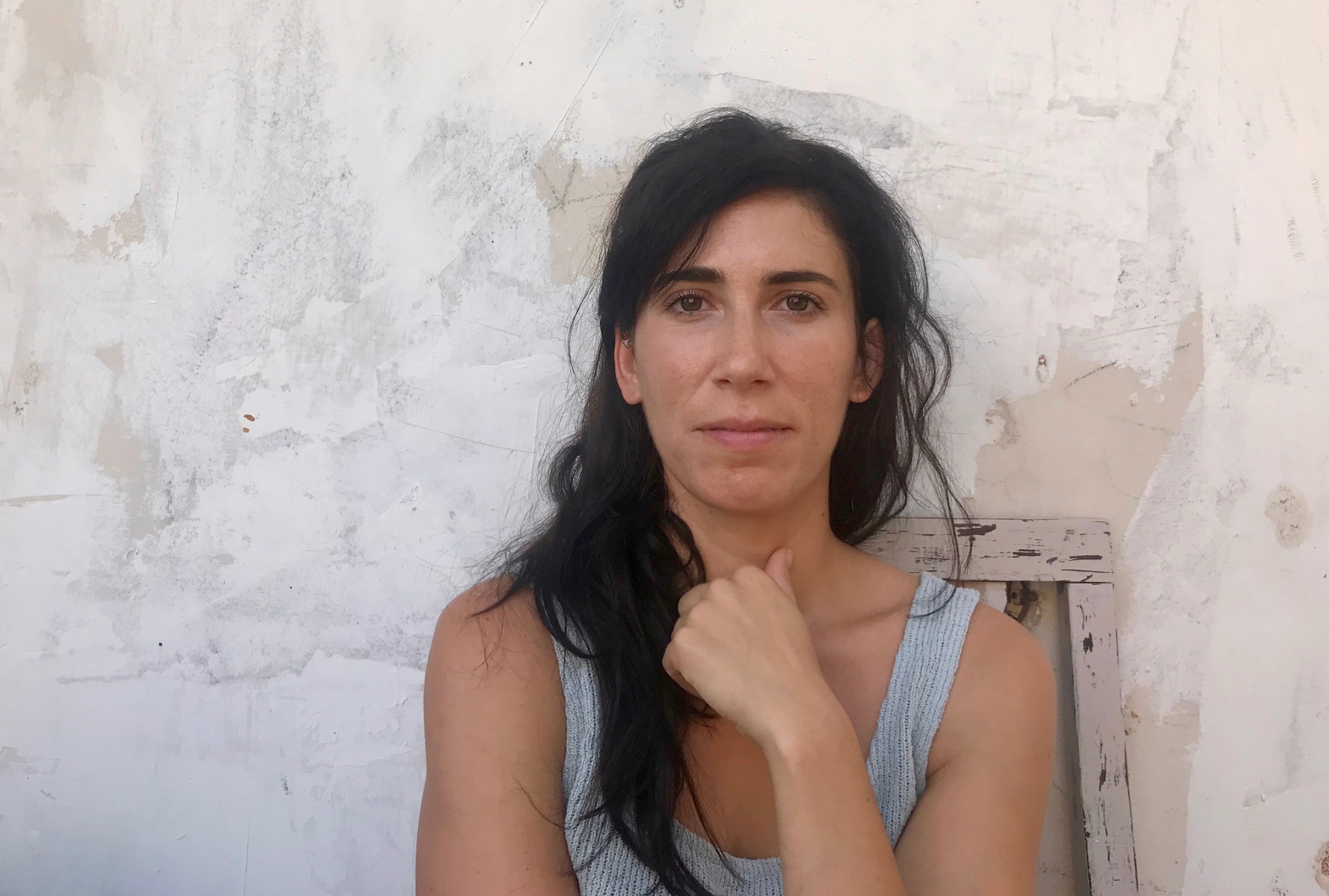 A portrait of a woman with long black hair, gently smiling, her hand resting in front of her neck. She is wearing a grey tank top against a white textured background.