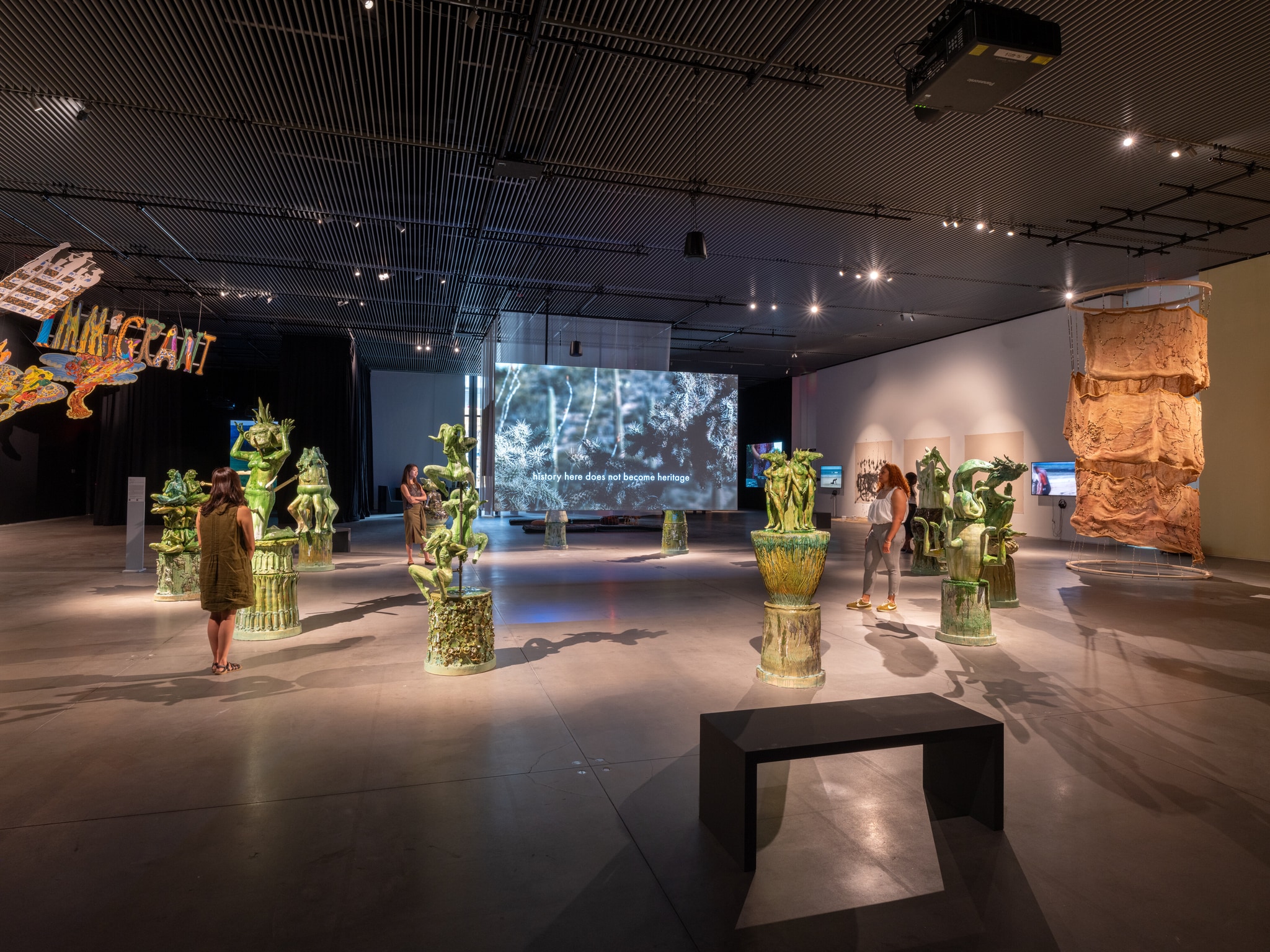 An installation view of a gallery exhibition. The gallery is a cavernous space without walls and cast in dim light. At the center stand several sculptural artworks including green ceramic sculptures of animals from the Eastern zodiac. Video screens with video artworks playing dot the space. 