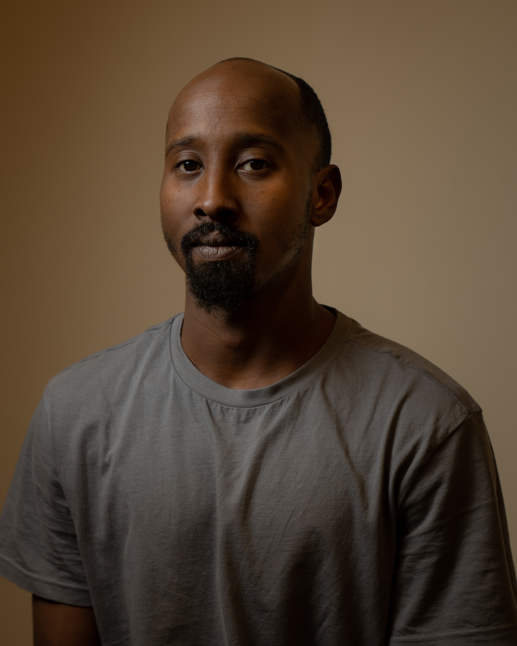 A portrait of Mohammed Smahneh against a neutral brown background. He wears a gray longsleeved shirt. He his bald and has a beard and he looks directly at us.