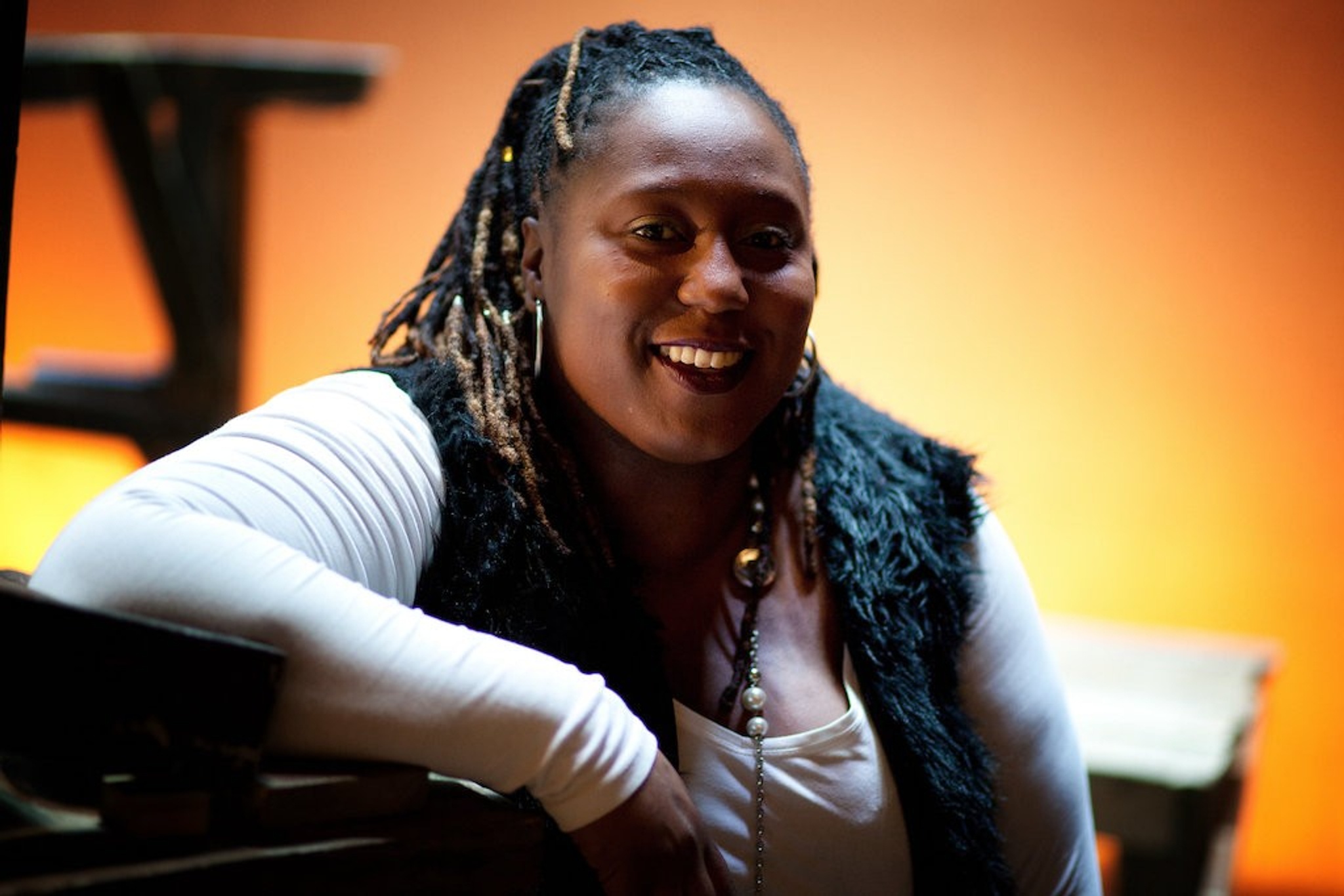 Nadine Lee sits comfortably while leaning on her right arm. She smiles against a background lit by soft orange light. She wears a black furry vest over a sand-colored long-sleeve shirt.